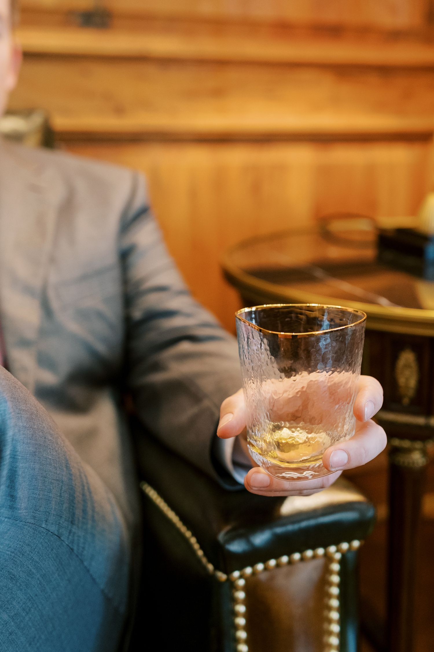 Glass of whiskey being held in hand against wooden bar background with dim lighting.