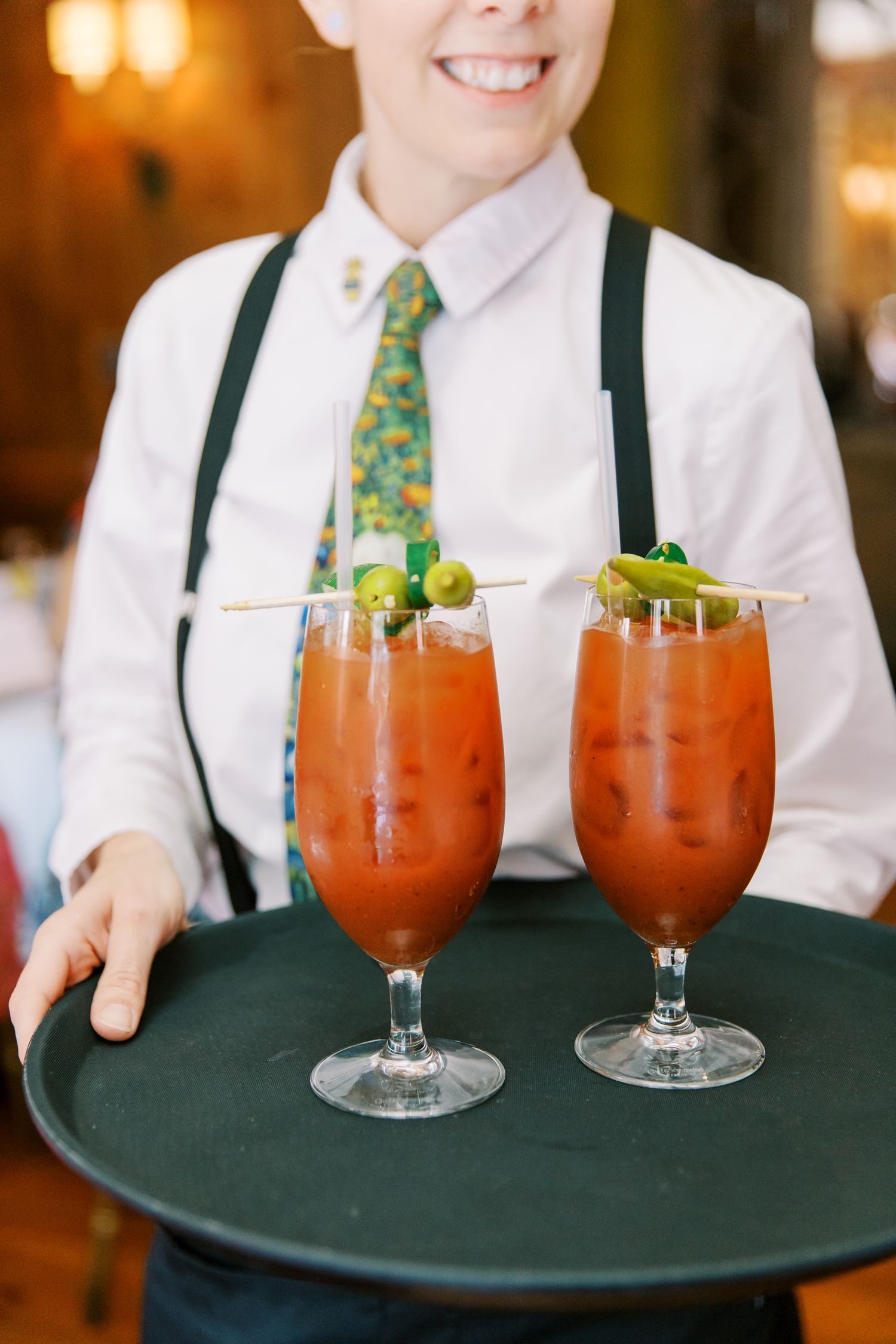 Server holding two garnished Bloody Mary cocktails on a serving tray in an elegant setting.