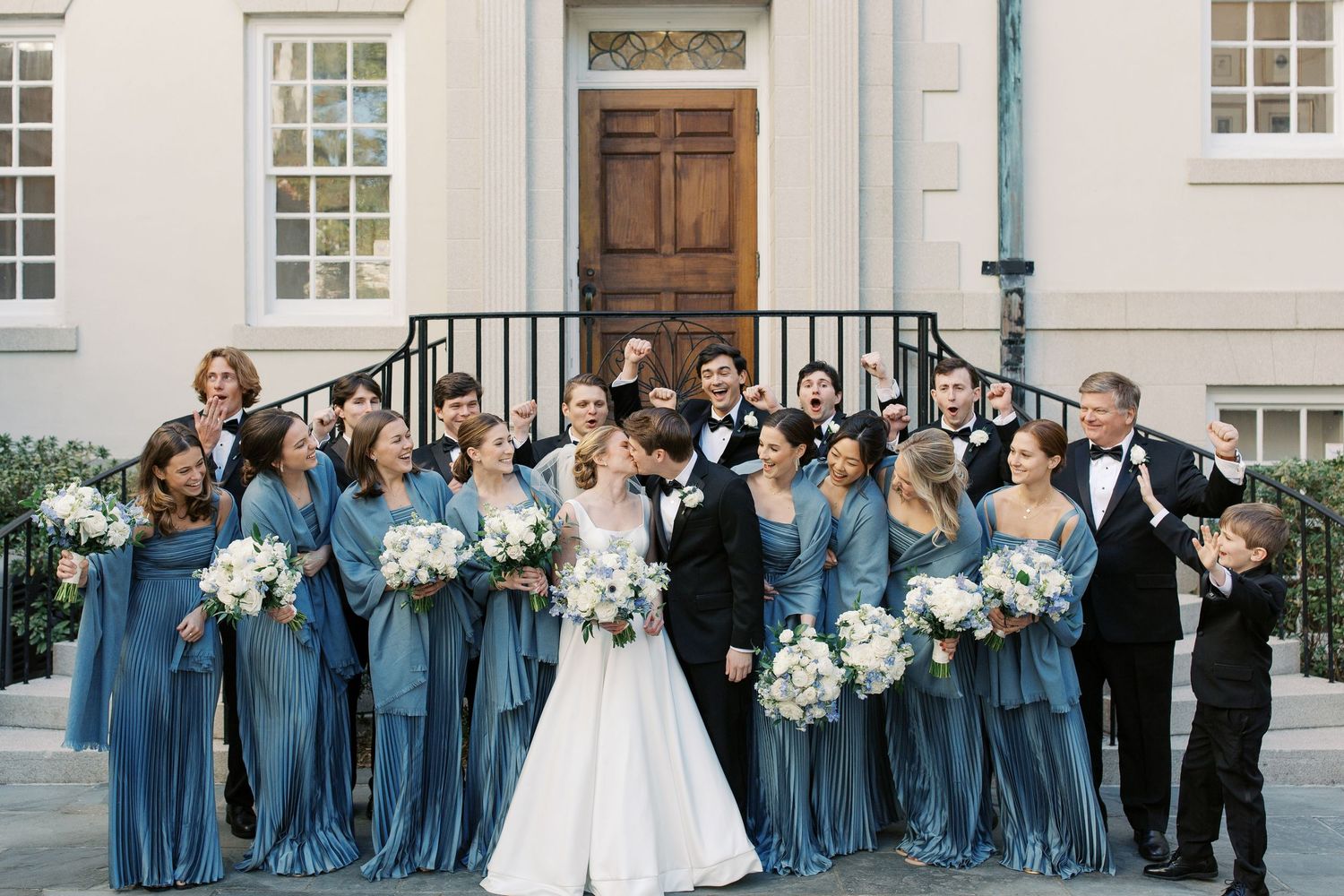 Wedding party in blue bridesmaid dresses and formal attire poses together outside a white stone building.
