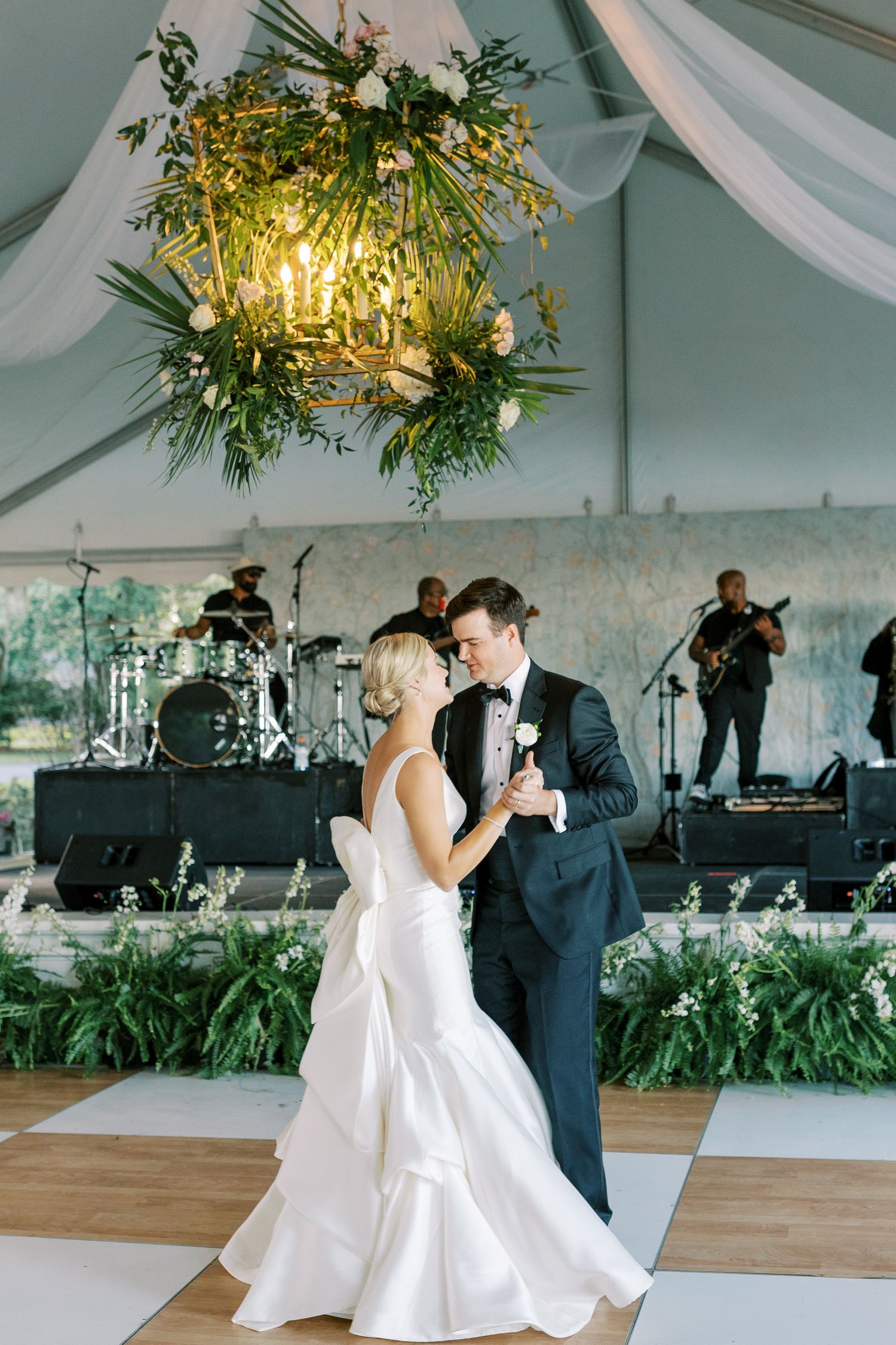 Couple shares romantic first dance under floral chandelier with live band performing on stage.