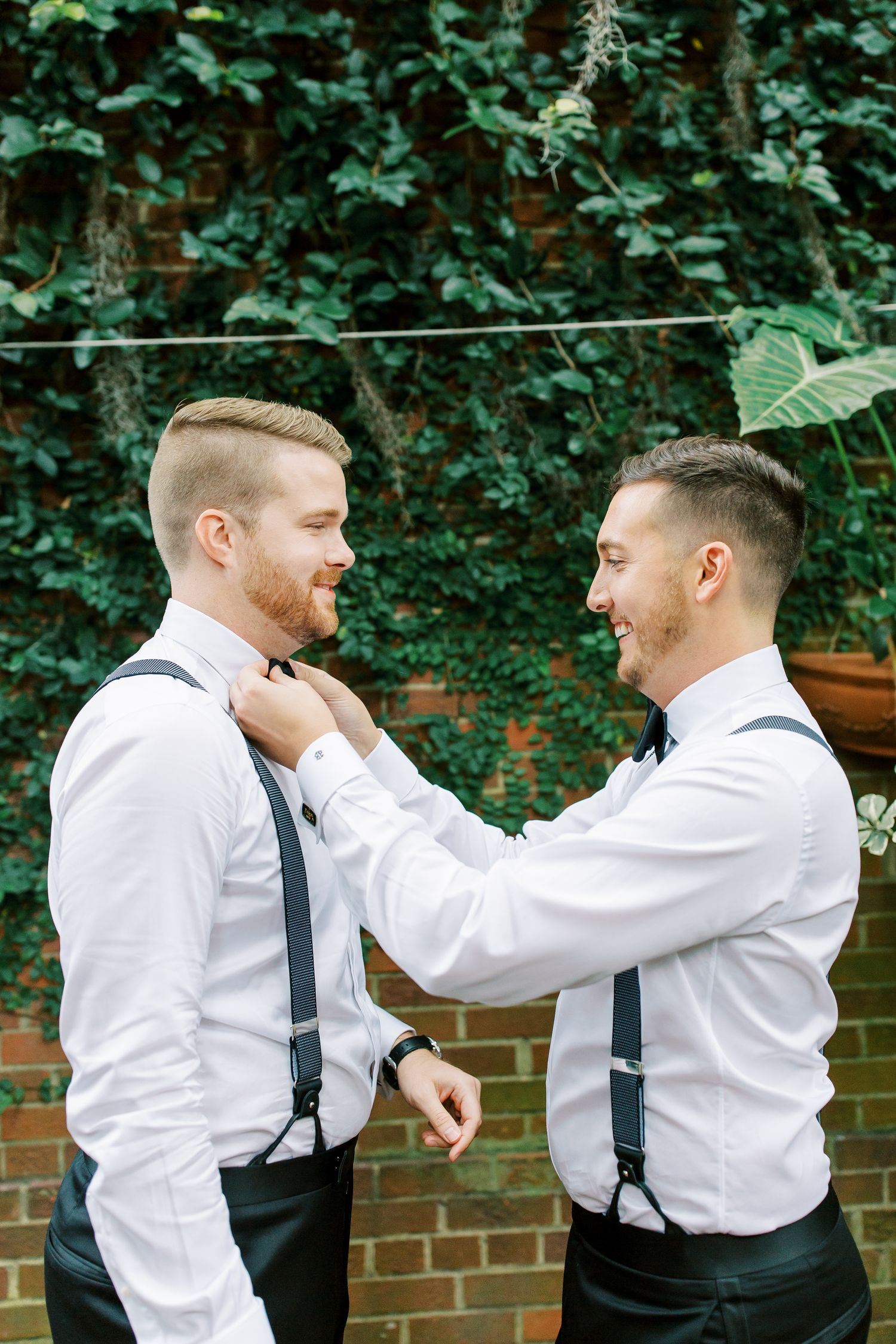 Two grooms adjusting each other's bow ties while wearing matching white dress shirts and suspenders in a garden setting.