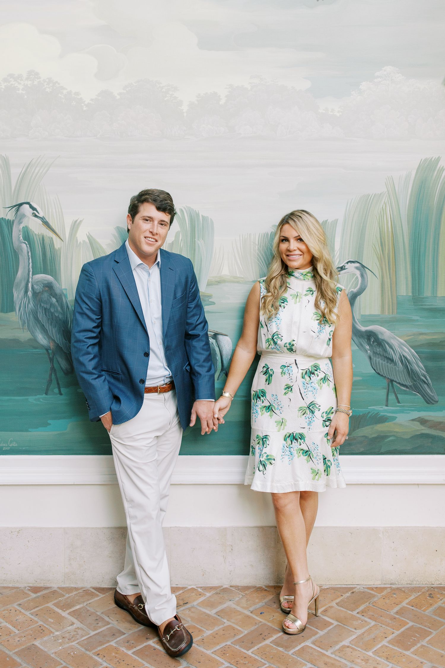 A couple poses in front of a scenic wallpaper mural featuring herons and water lilies in muted blue-green tones.