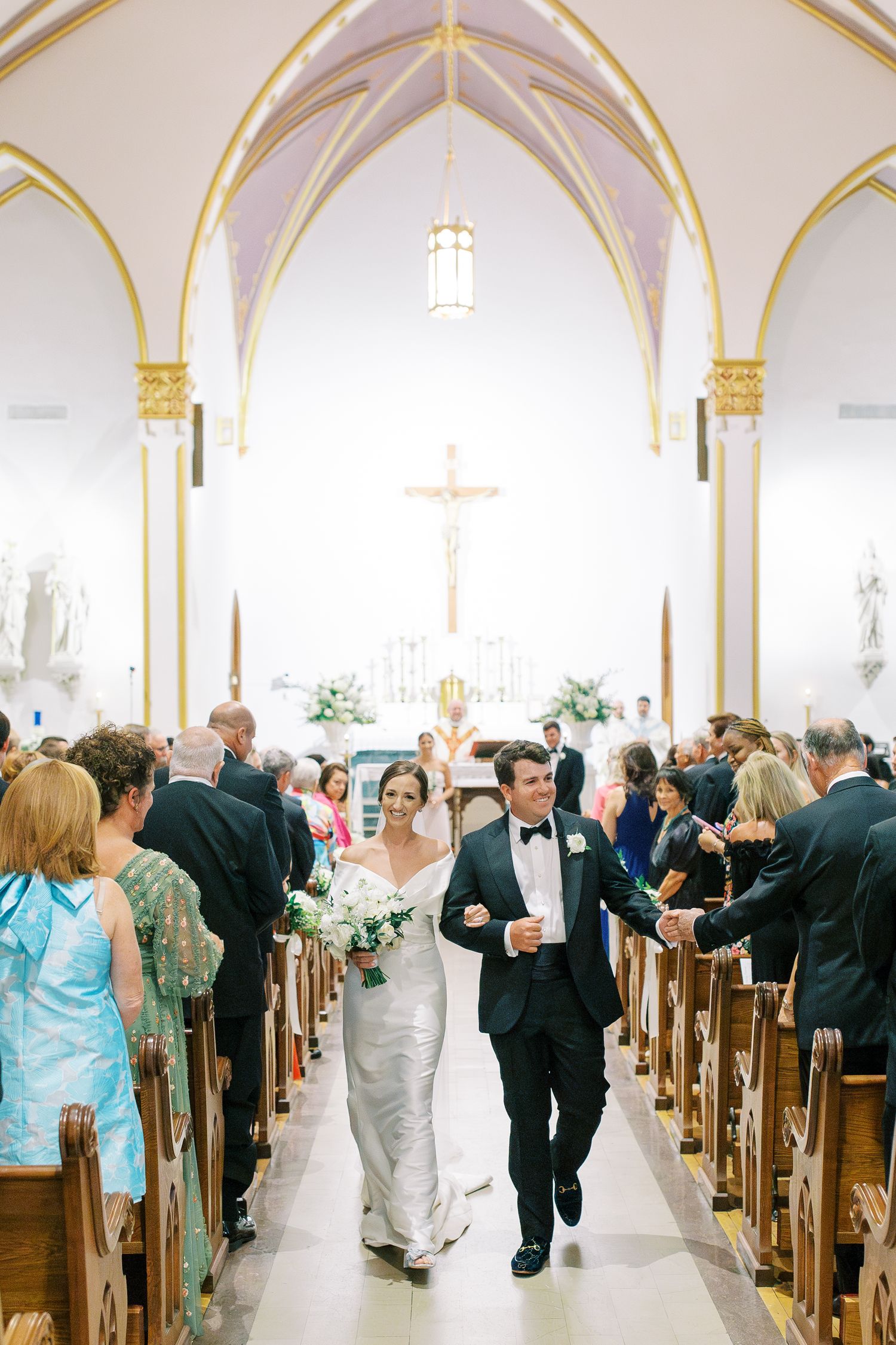 Newly married couple exits church ceremony through aisle lined with guests beneath gothic arches and golden trim at Scared Heart Church.