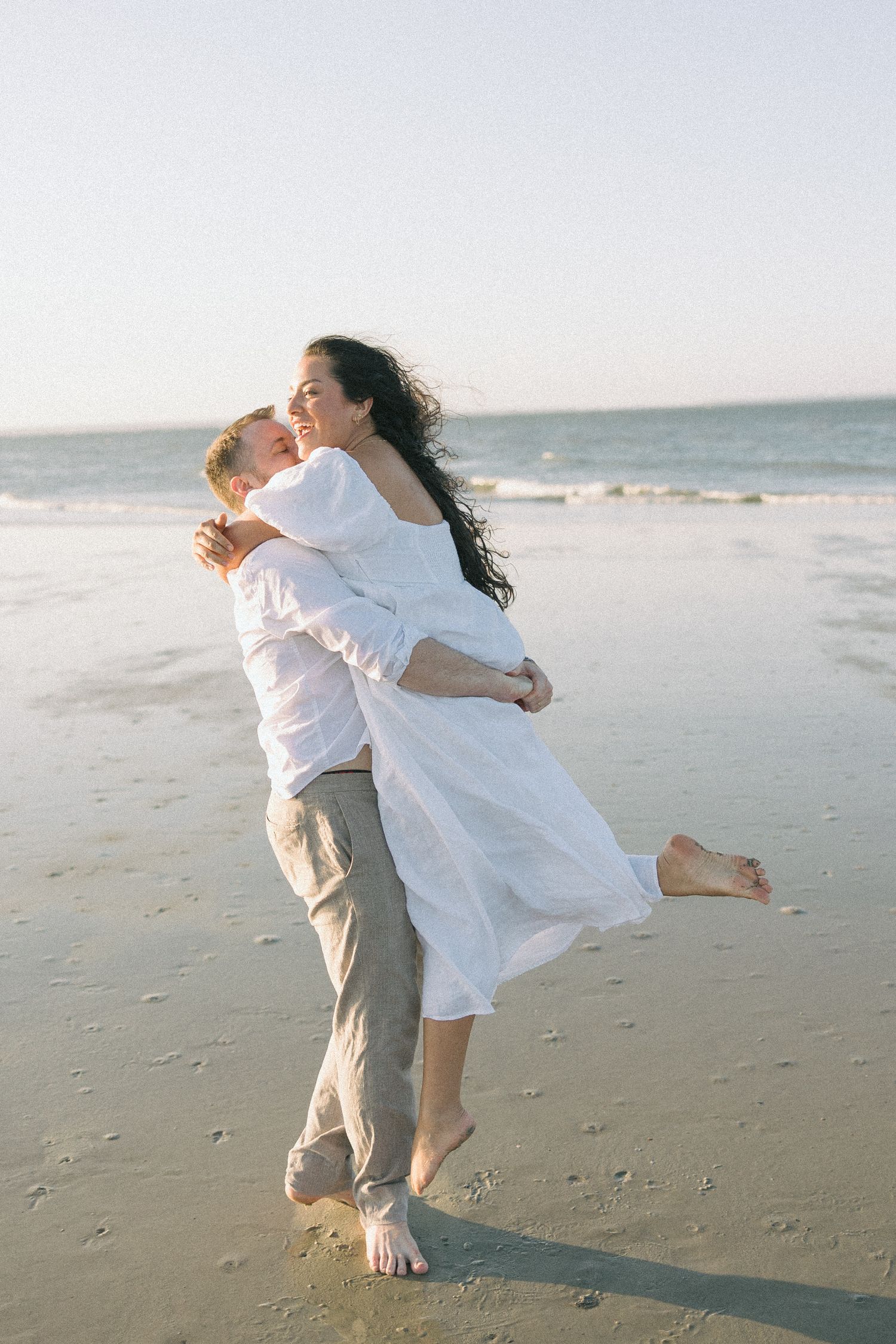 A couple in white clothing dances joyfully together on a sandy beach at sunset.