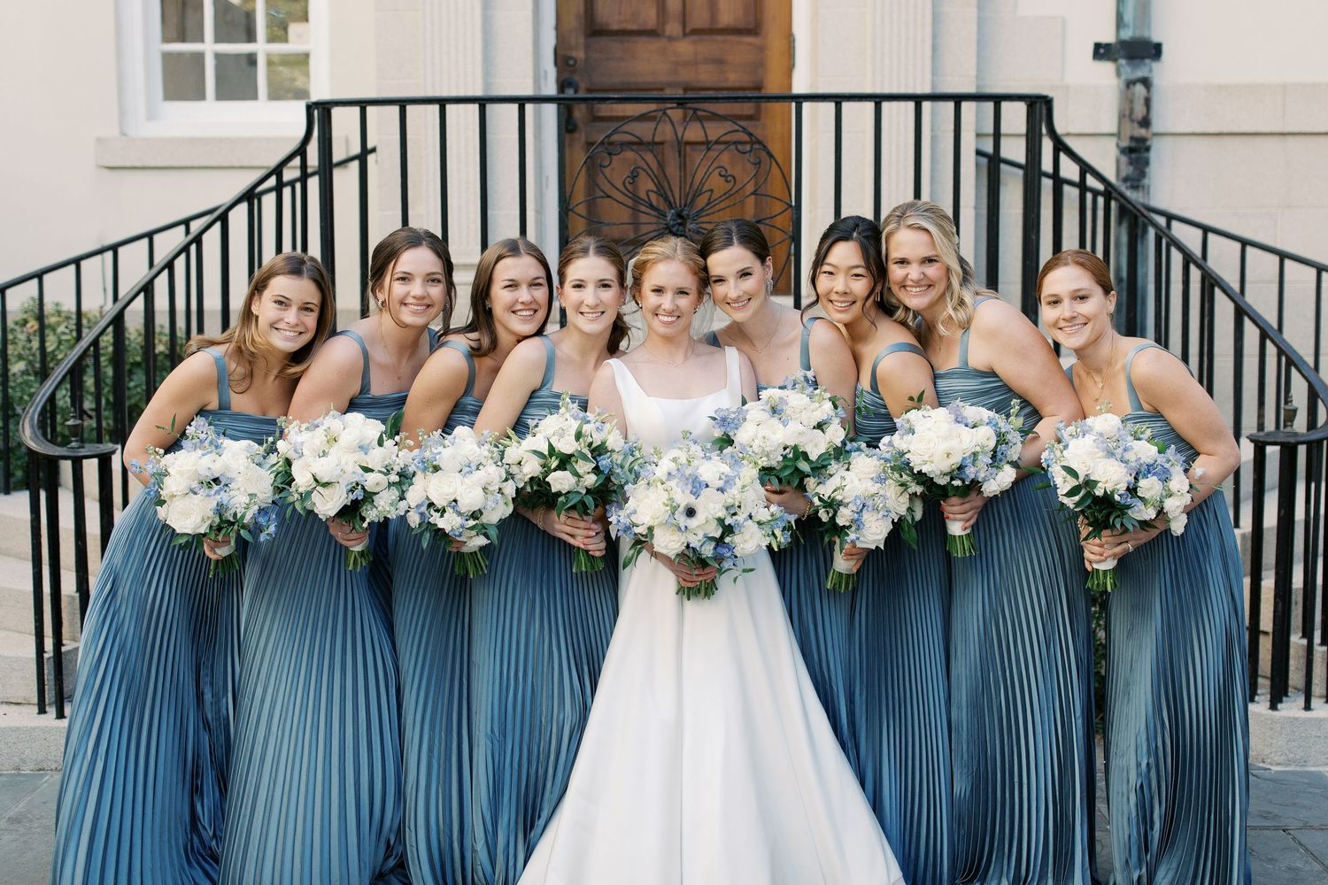 Wedding party lined up outside church wearing blue bridesmaid dresses and holding bouquets.