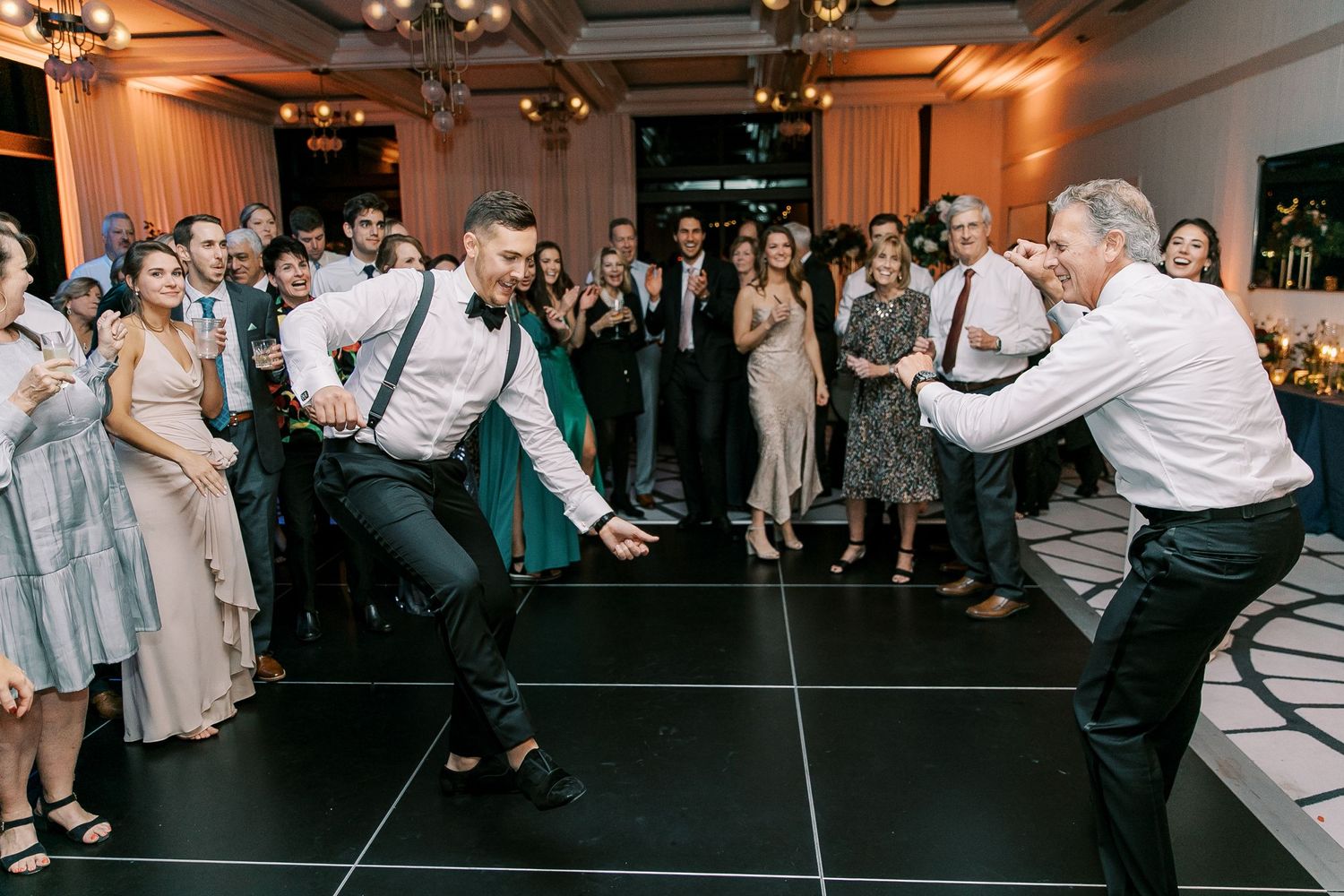 Groom and dad in suspenders dance enthusiastically on a dark dance floor while wedding guests watch from the sidelines.