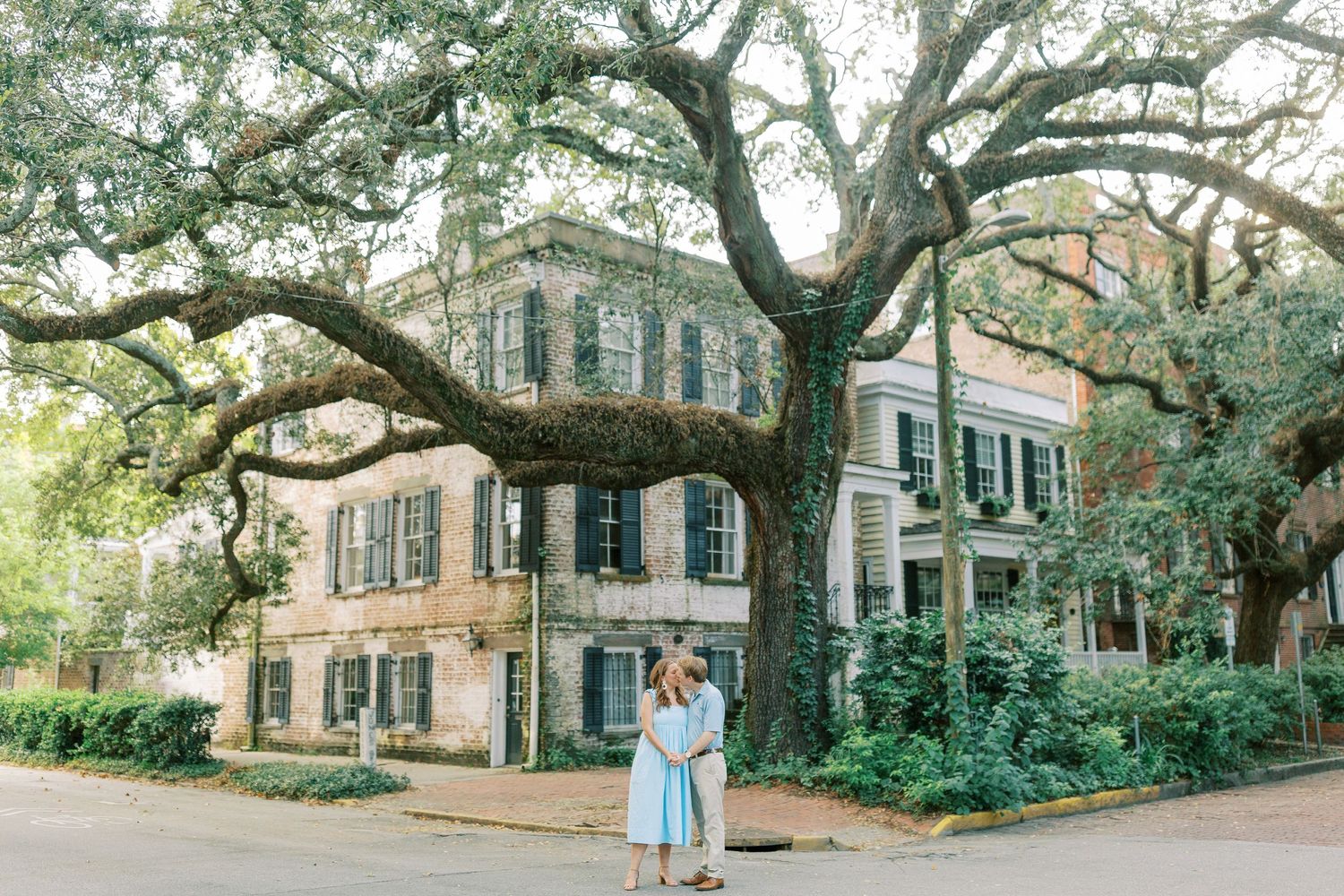 A romantic couple stands beneath sprawling oak trees in front of a historic Savannah mansion with blue shutters.