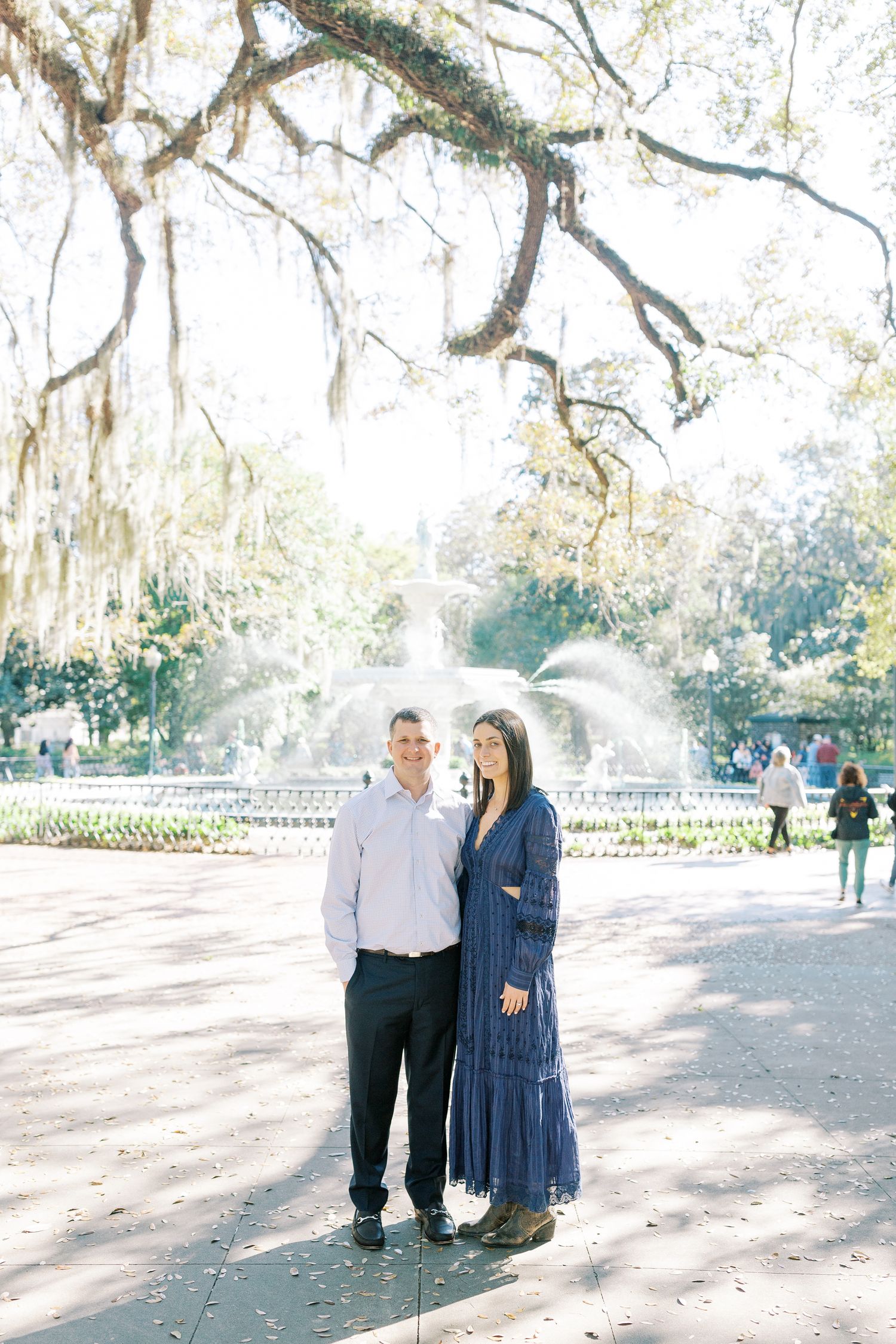A couple stands together under moss-draped oak trees in a sunlit park with the Forsyth Park fountain in the background.