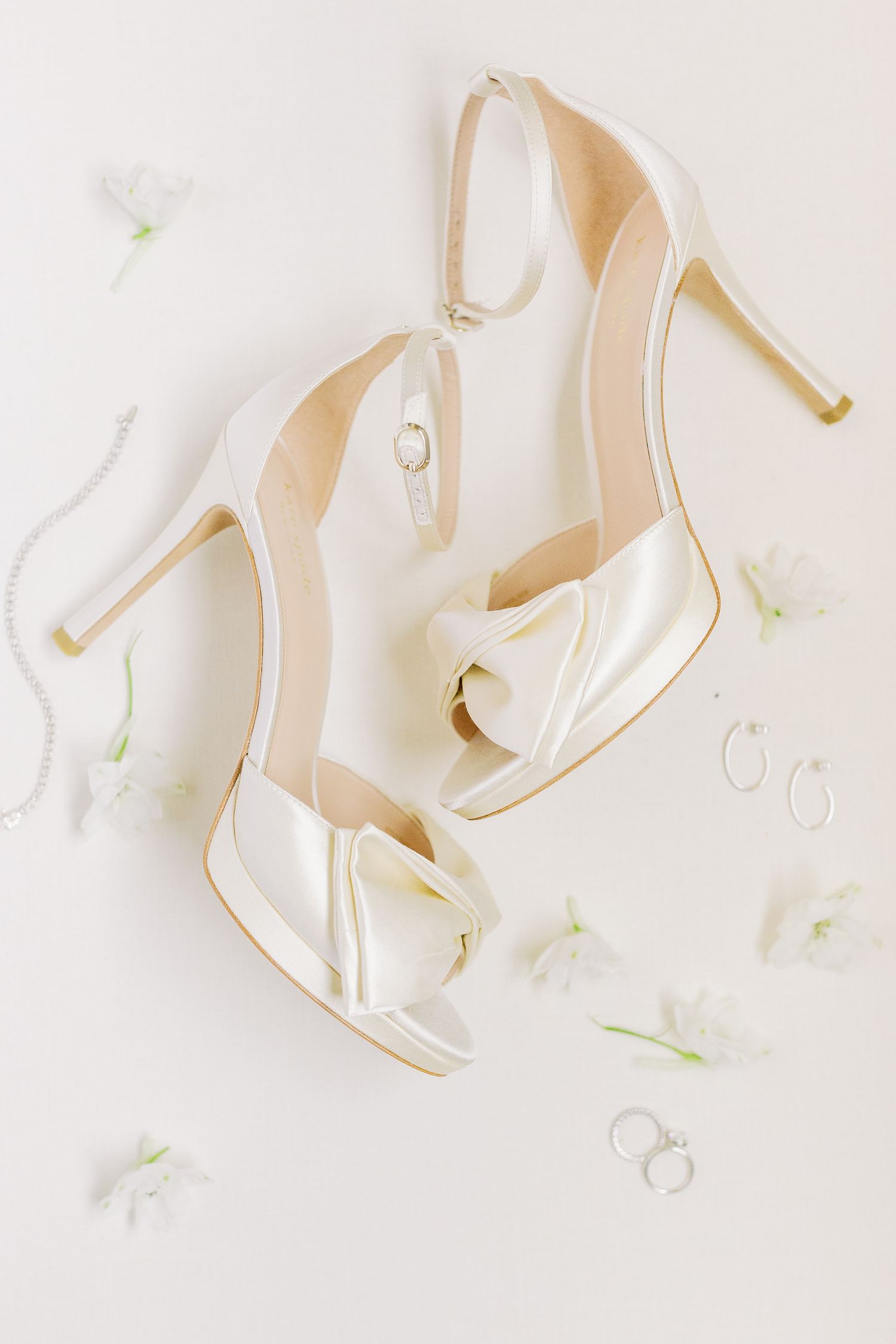Elegant white satin peep-toe wedding heels with small floral accents photographed on light background.