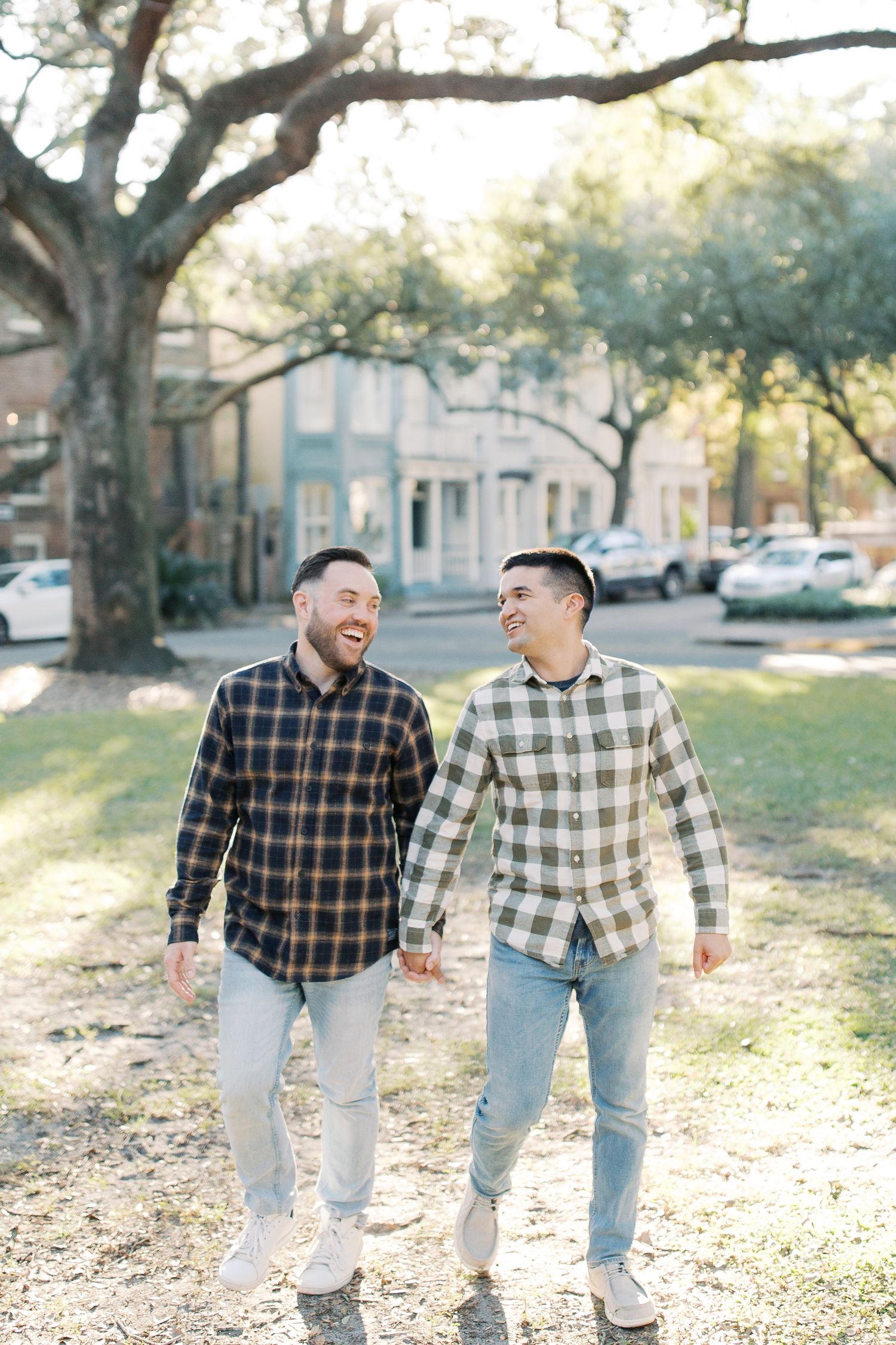Joyful couple walking hand in hand under sunlit oak trees in matching casual attire.