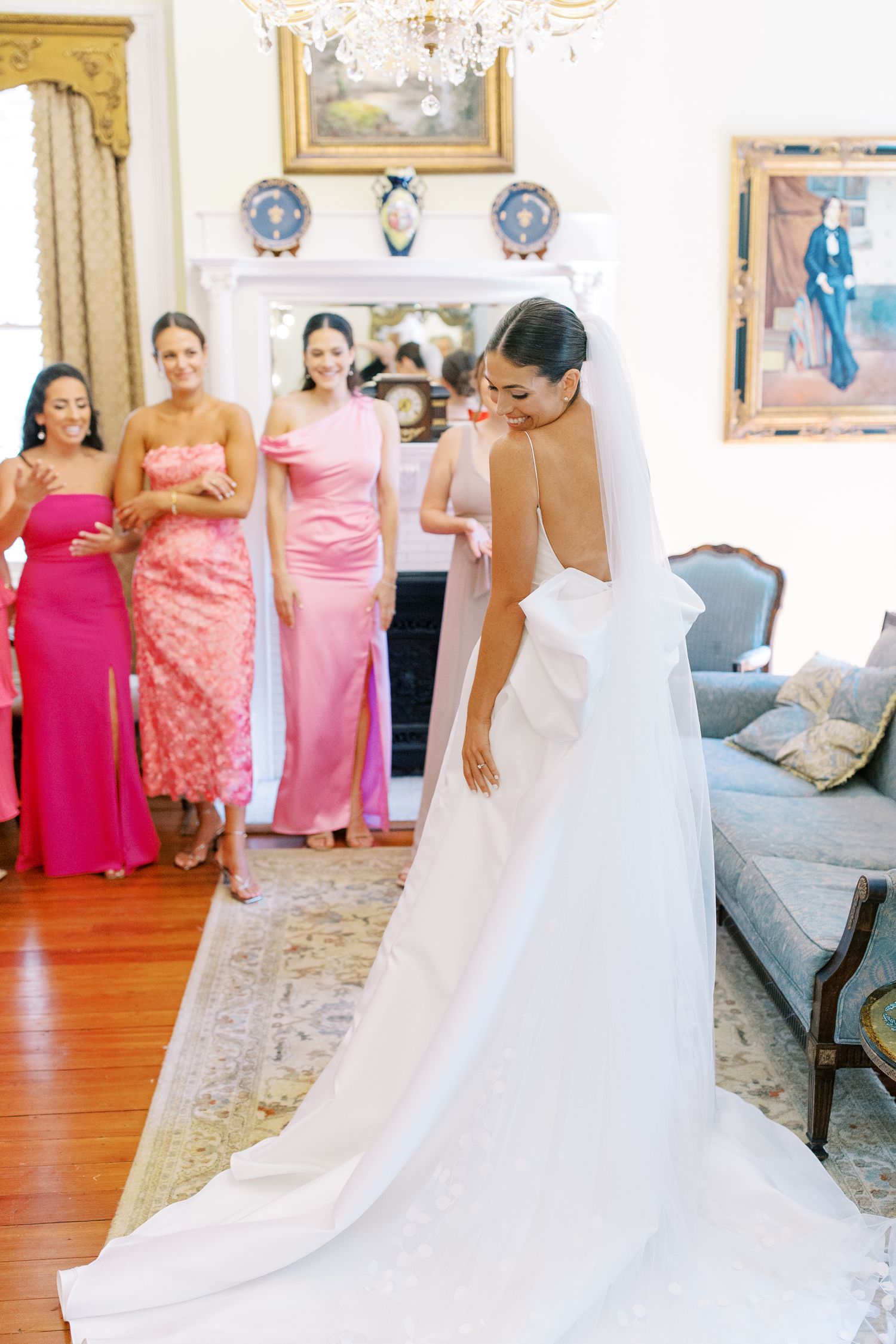 A bride in a flowing white gown shows off her dress to attendants in pink dresses.