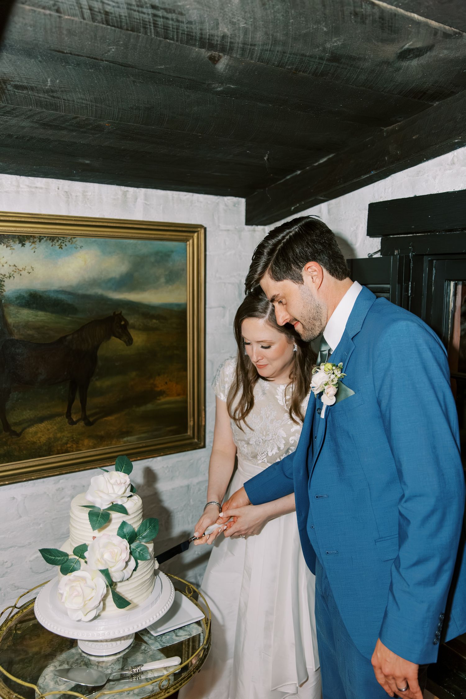 Newlyweds in blue suit and white dress cutting their wedding cake together in a rustic indoor setting.