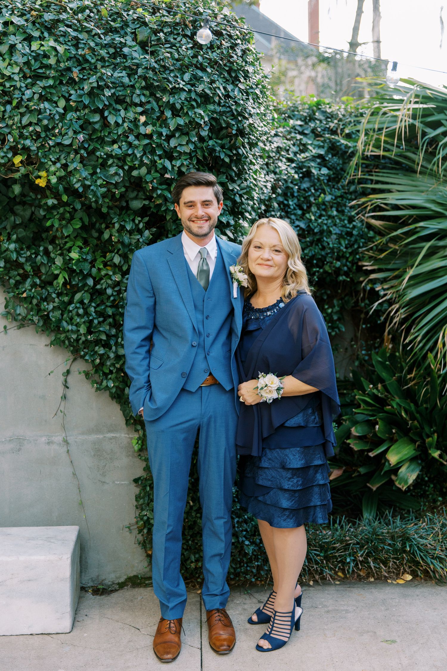 A couple poses together against a lush green hedge backdrop wearing coordinating navy blue outfits.