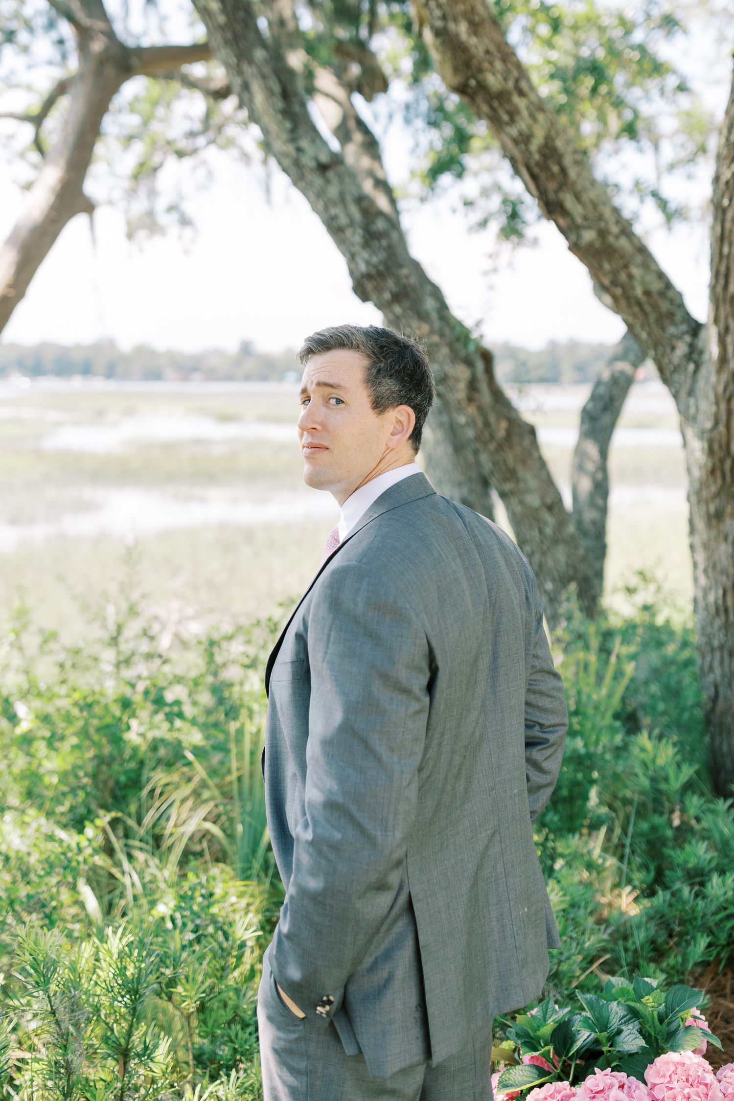 Groom in grey suit poses thoughtfully among trees and flowers in an outdoor garden setting.