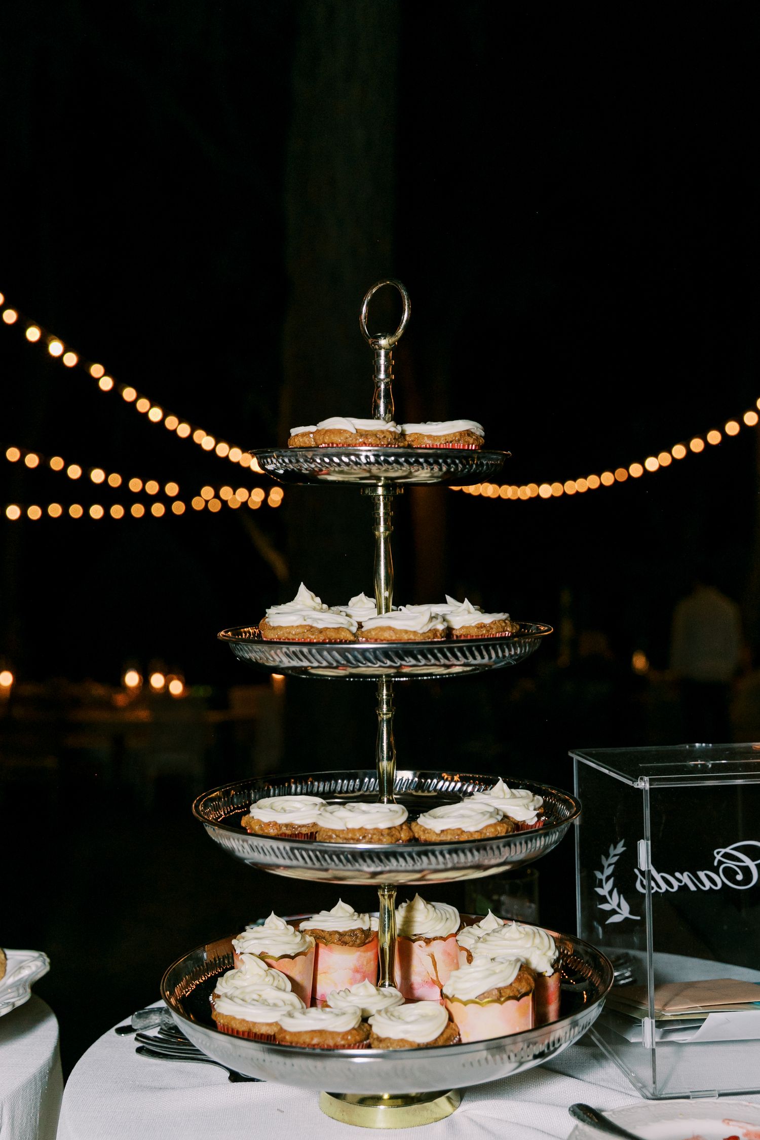 A tiered dessert display with white treats on elegant silver platters under string lights.