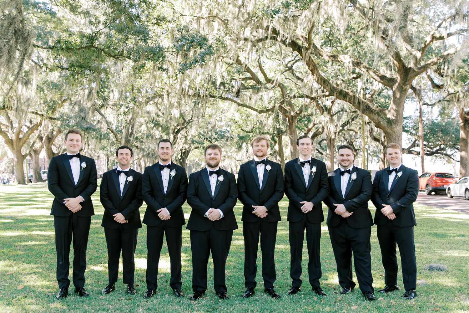 A line of groomsmen in black tuxedos stand under elegant oak trees draped with Spanish moss on a sunny day.