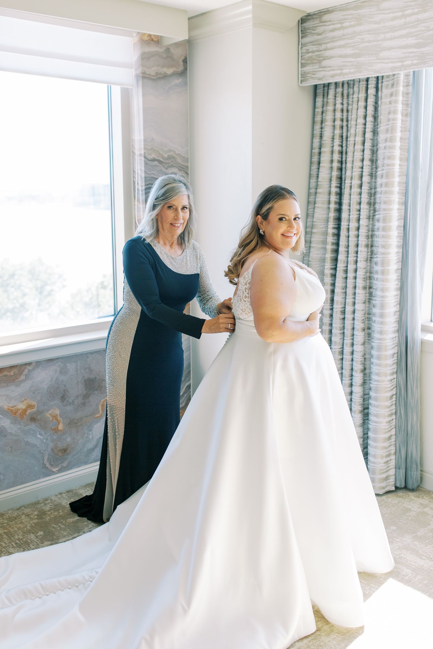 A bride in a classic white ballgown receives help getting dressed near a bright window.