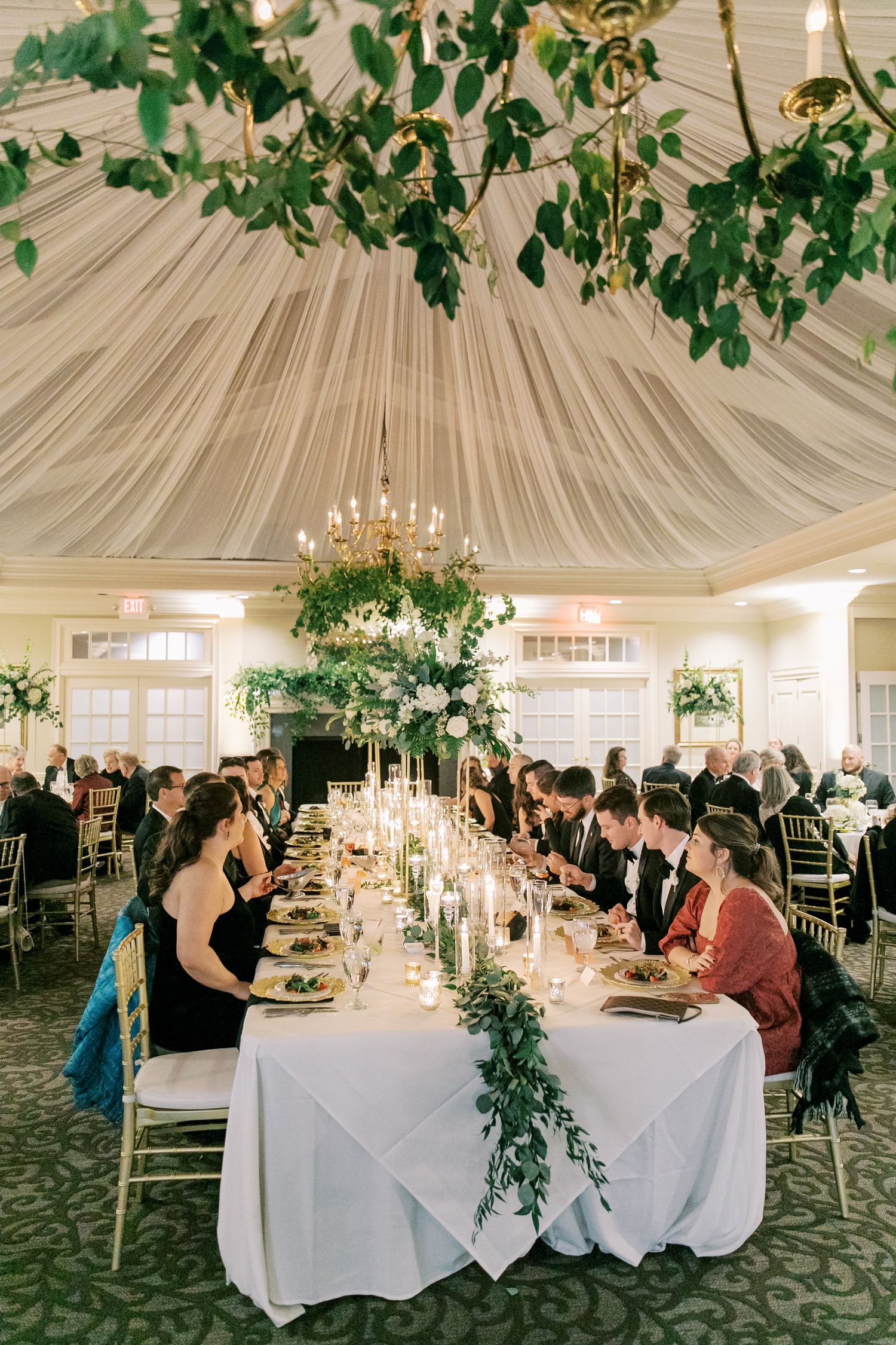 Elegant wedding reception with draped ceiling, chandelier and long dining tables adorned with greenery and candles.
