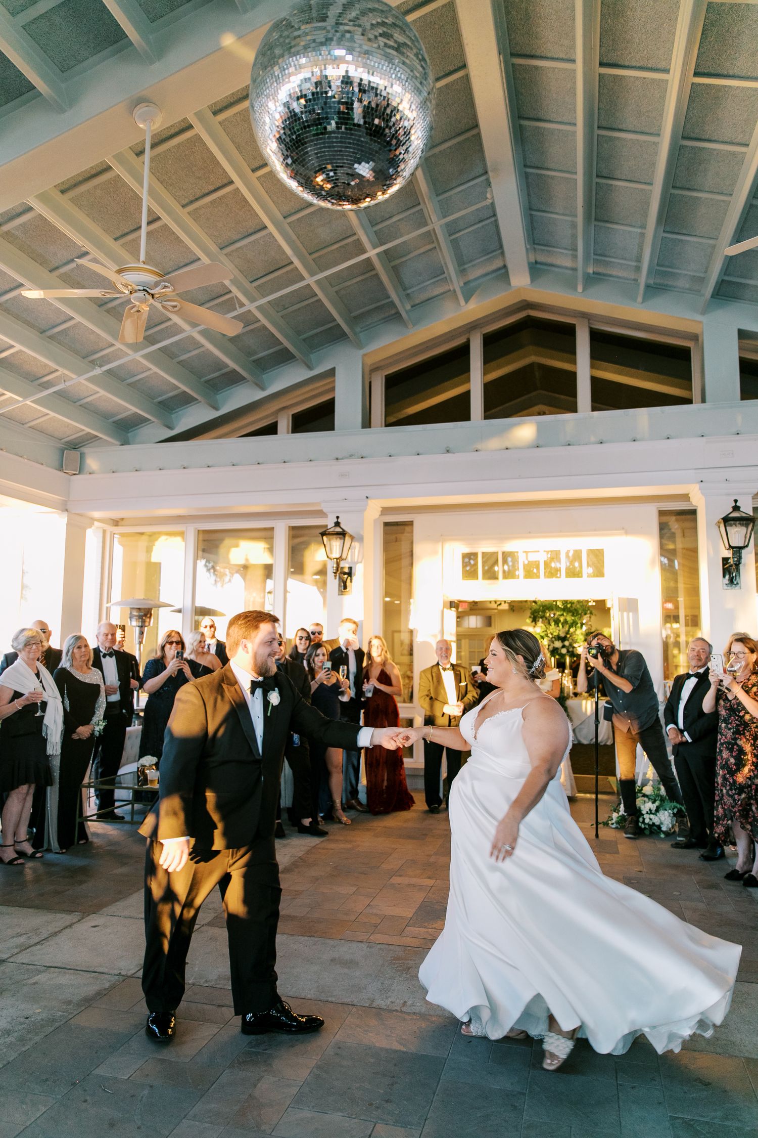 Wedding guests gather under a sparkling disco ball to watch a couple's first dance in an elegant reception hall.