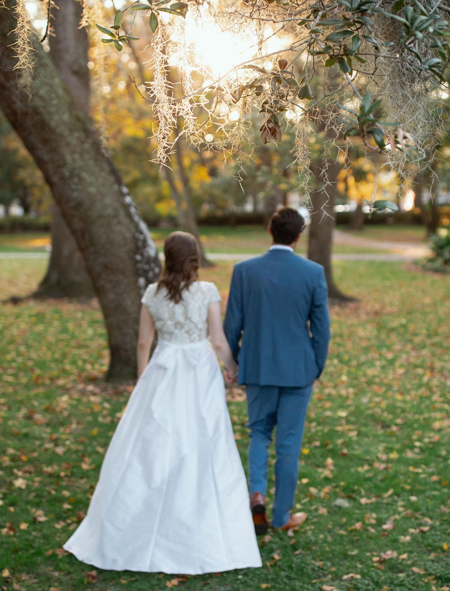 Couple walks through a sunlit garden with autumn leaves scattered on the ground during their wedding day.