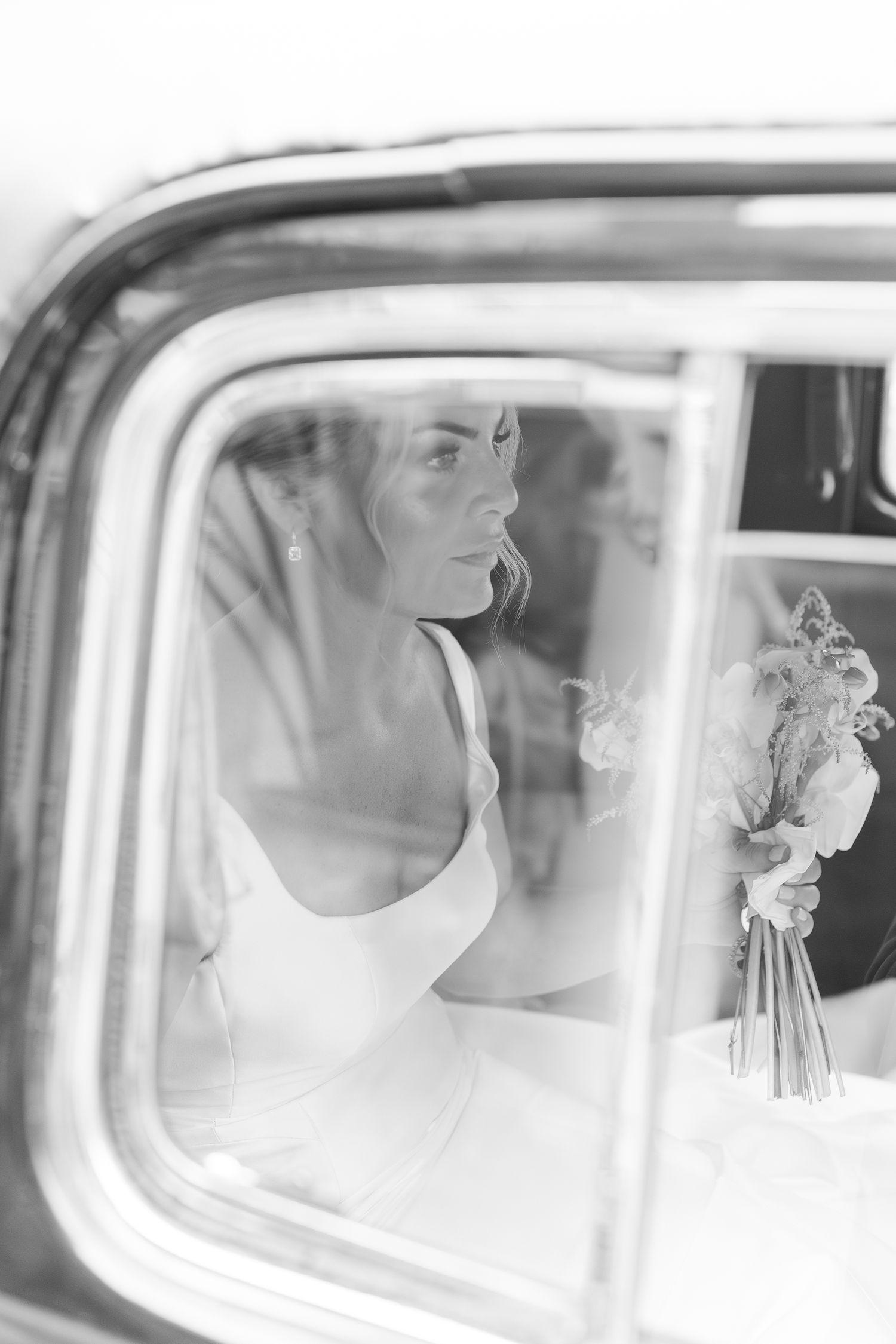 Artistic black and white photo of a bride through a vintage car window holding a bouquet.