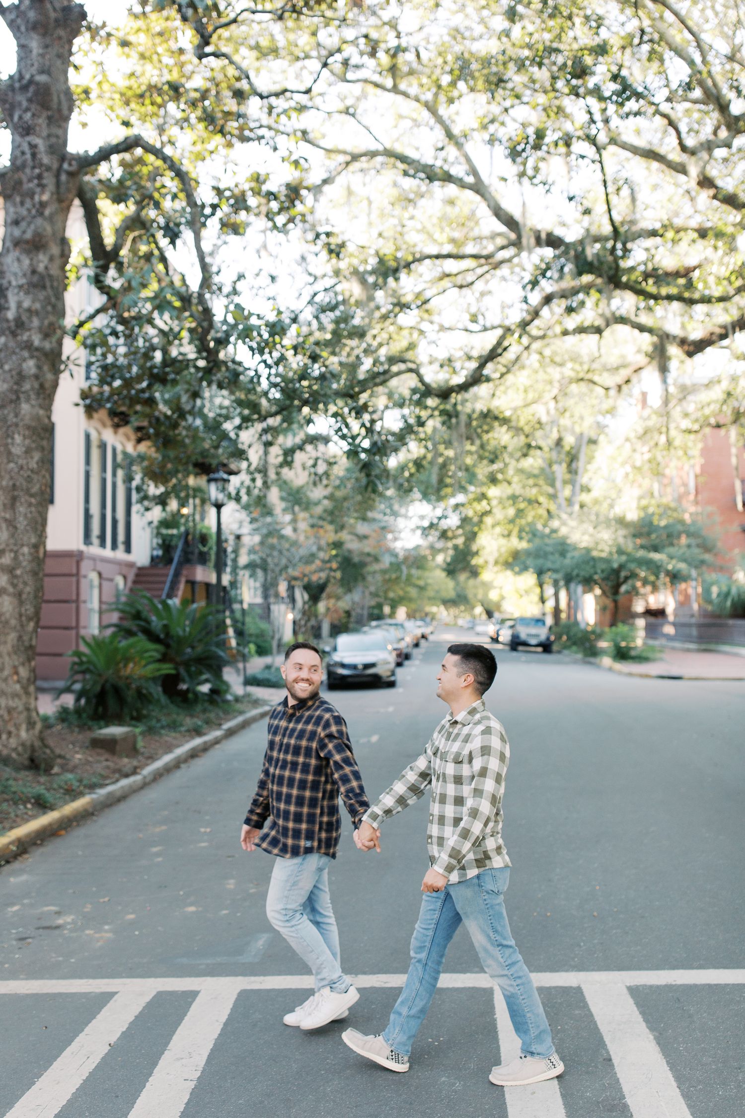 Two people crossing street in casual plaid shirts and jeans under tree canopy.