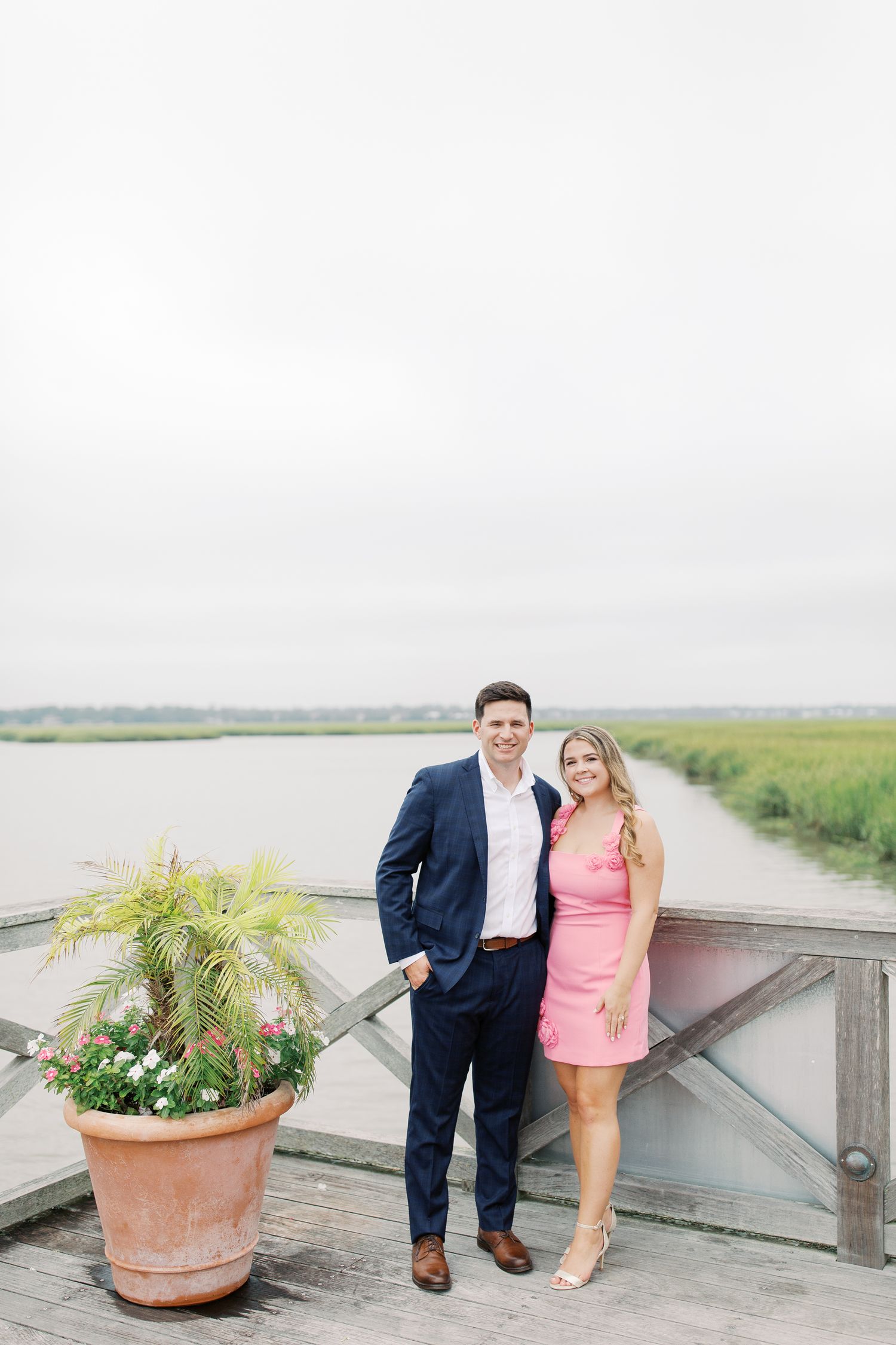 Couple standing on wooden deck overlooking marsh with potted flowers.