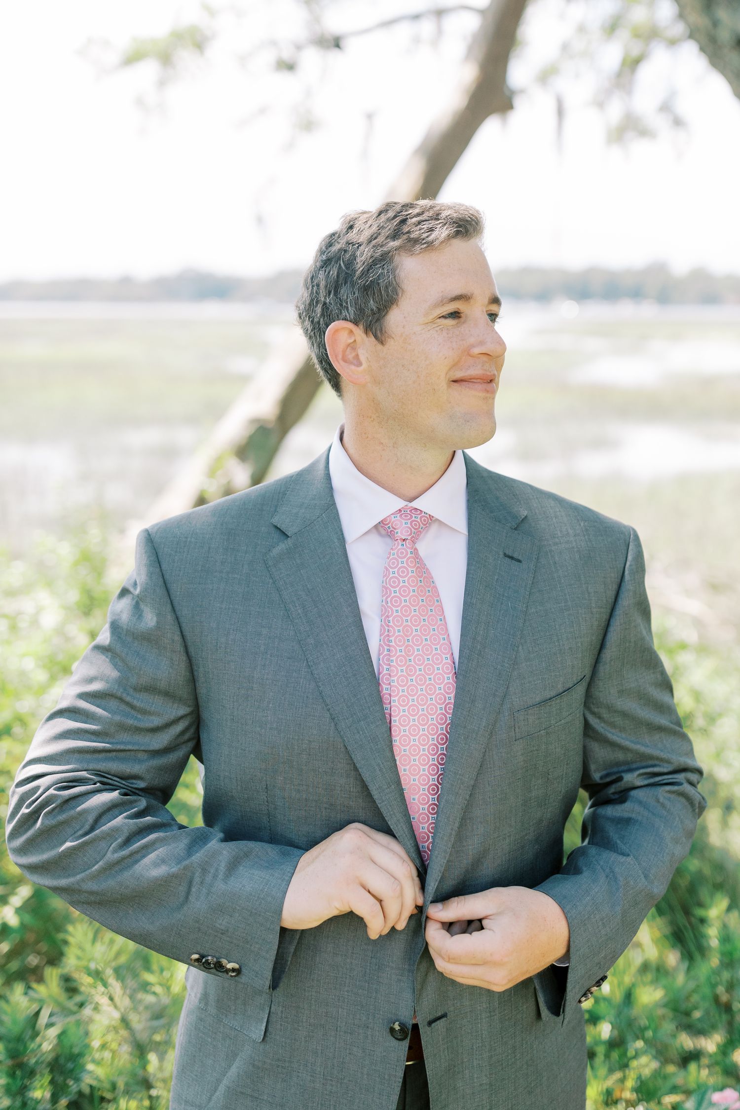 Groom in gray suit and pink tie smiles while standing outdoors near trees.