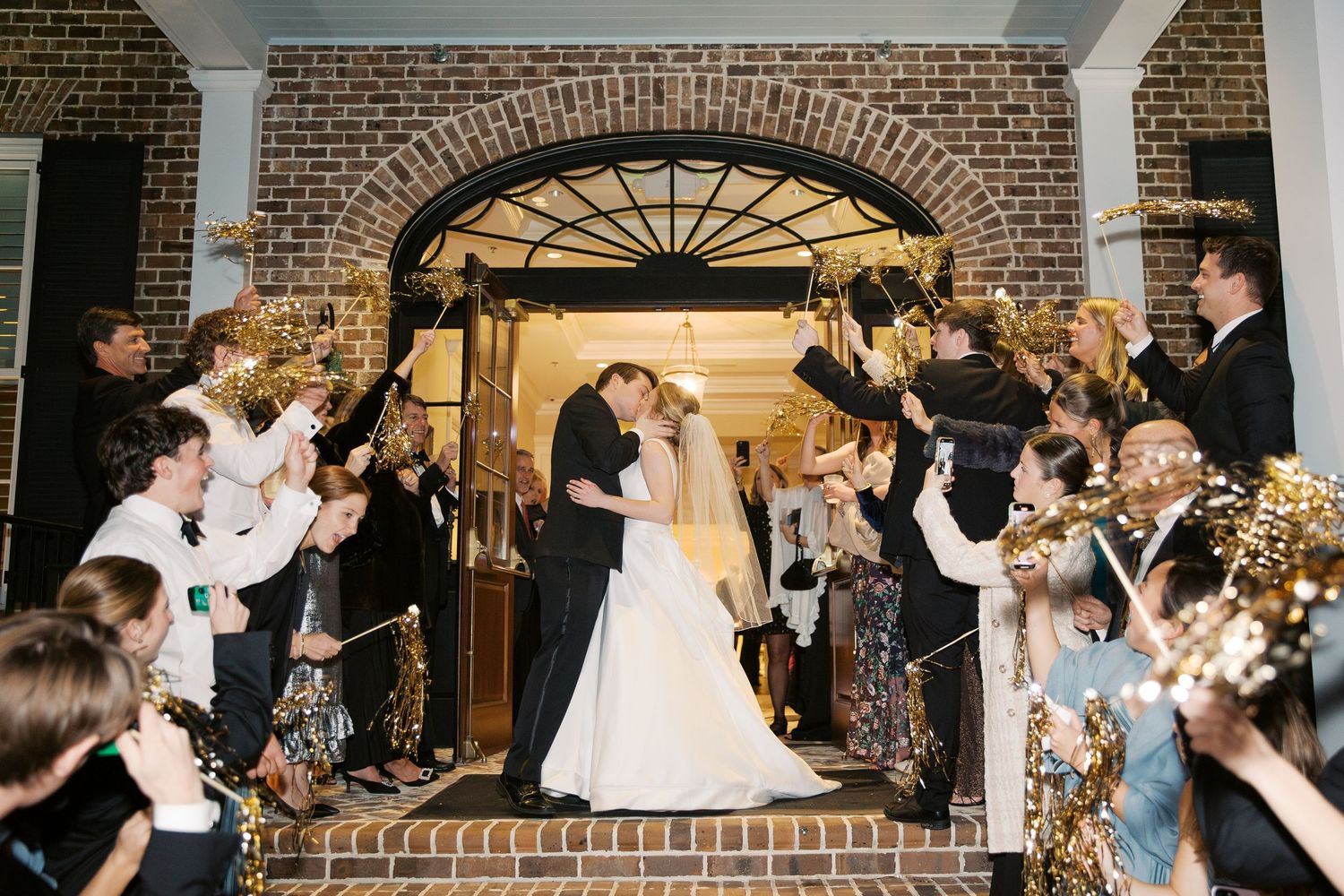 Wedding couple kissing under brick archway while guests celebrate with sparklers.