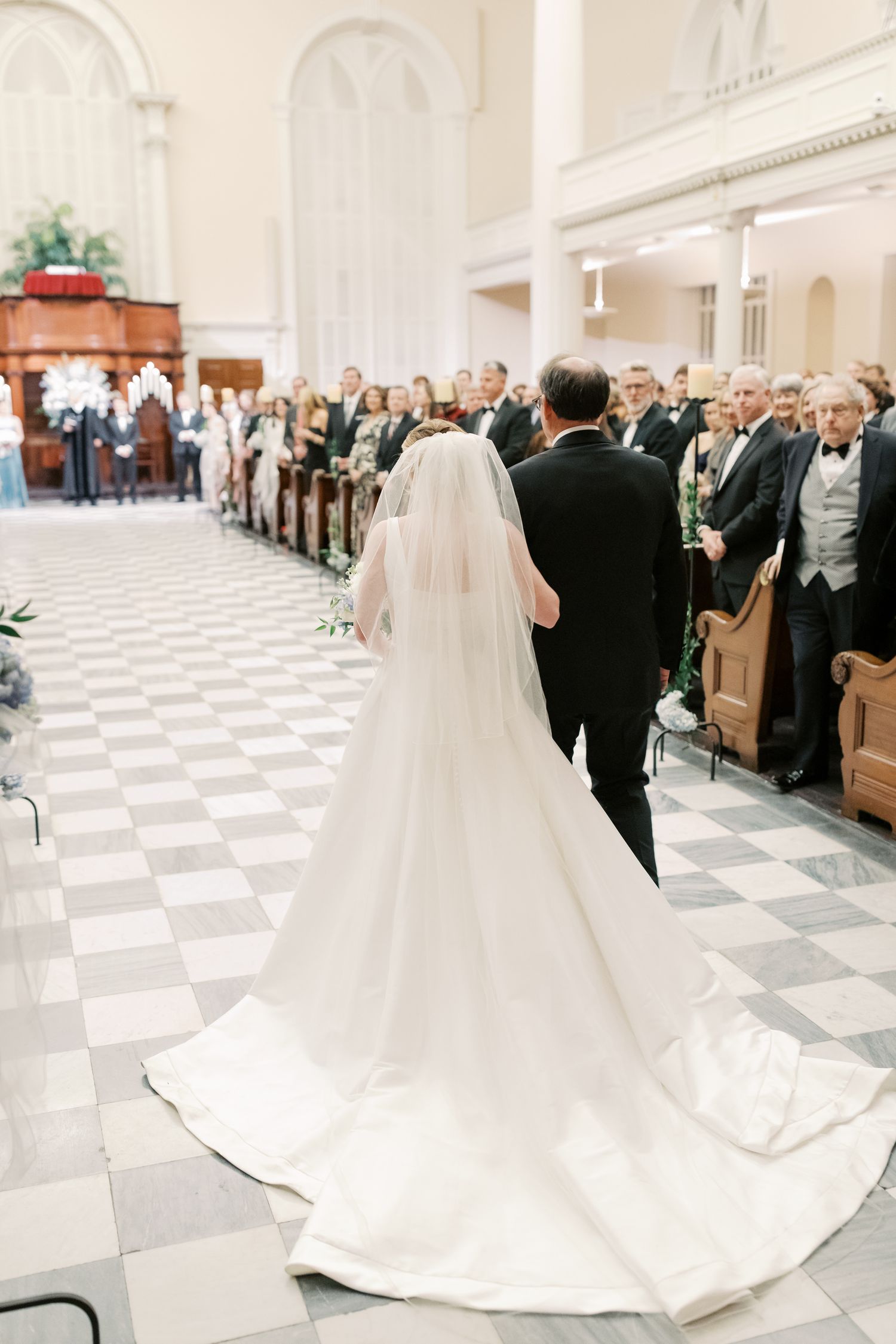 Church ceremony with long train of white wedding dress against checkered floor.