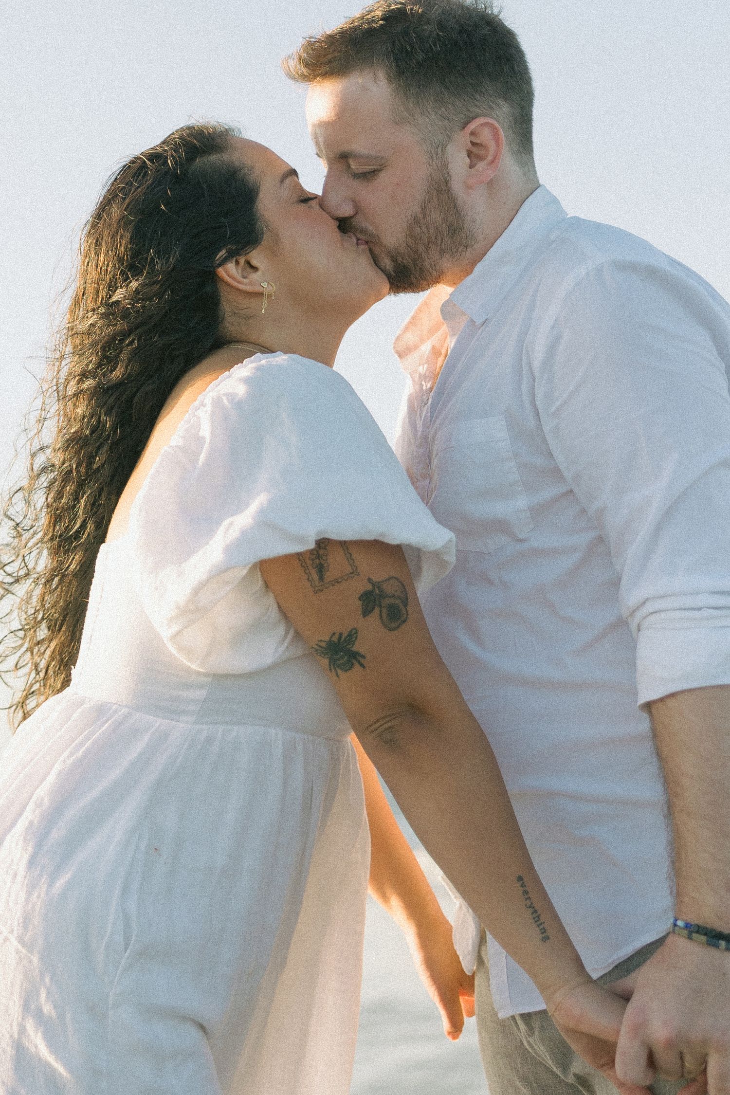 A romantic couple in white clothing share a tender kiss in soft sunlight, with visible tattoos on their arms.