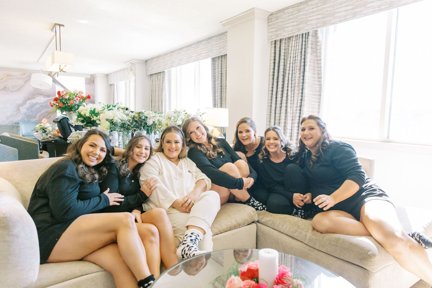 A group relaxes together on a beige sectional couch in a bright living room with floral decorations.