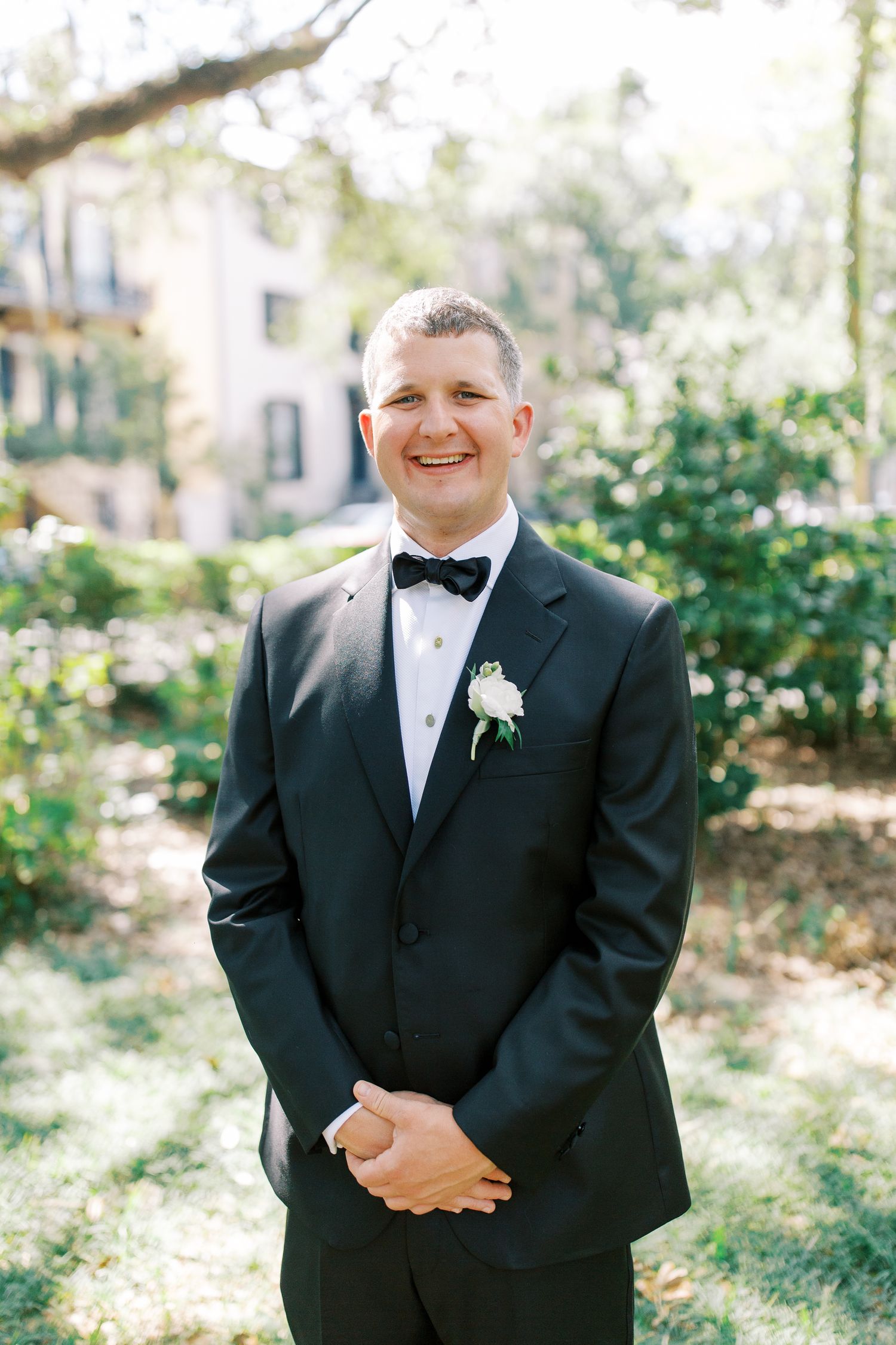 A groom in classic black tuxedo smiles in a garden setting.