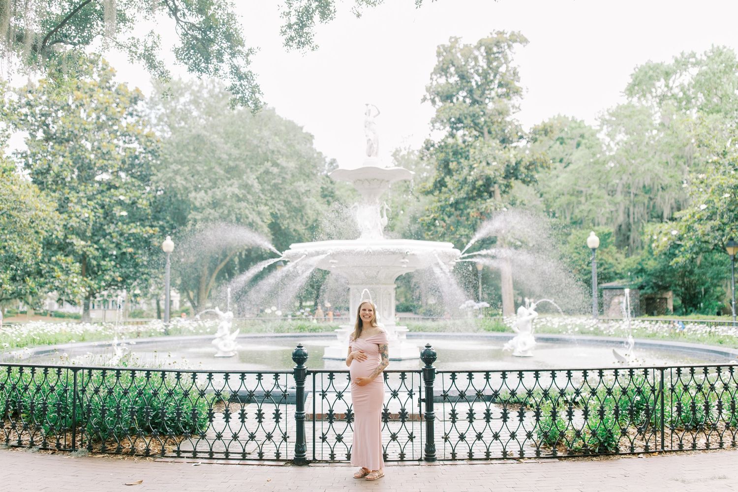 Forsyth Park fountain in a historic garden setting with ornate iron railings and lush greenery with pregnant mother in front.