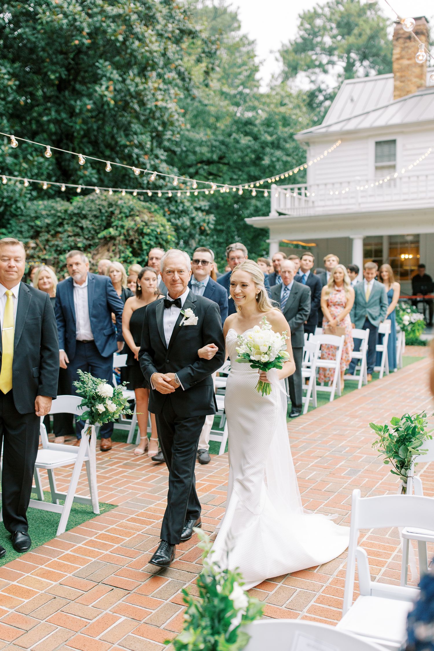 Outdoor wedding ceremony procession on brick pathway with string lights and white chairs.