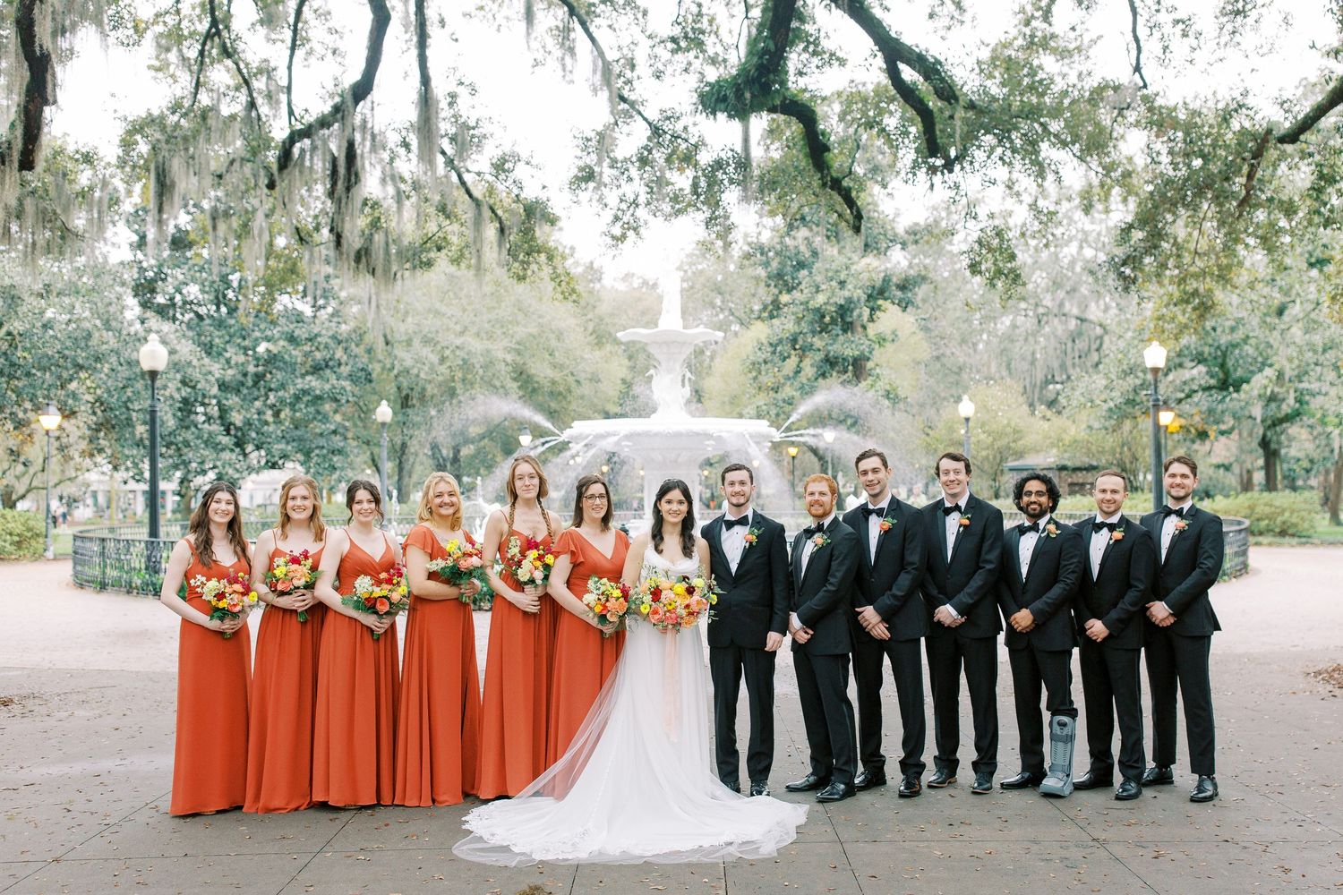 A wedding party poses outdoors with bridesmaids in orange dresses and groomsmen in black tuxedos in Forsyth Park.