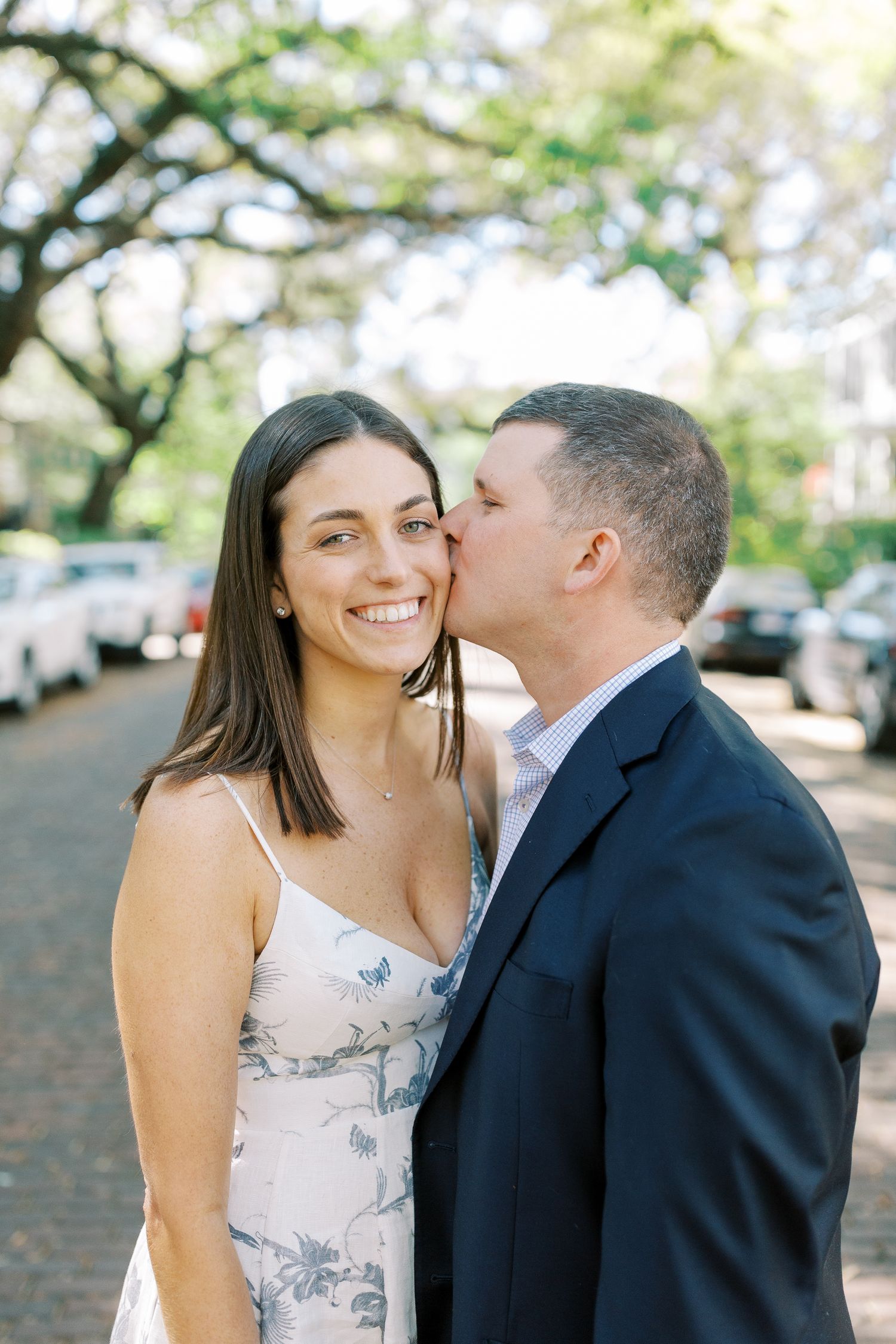 A couple shares a romantic moment under trees on a city street, with her in a floral dress and him in a dark suit.