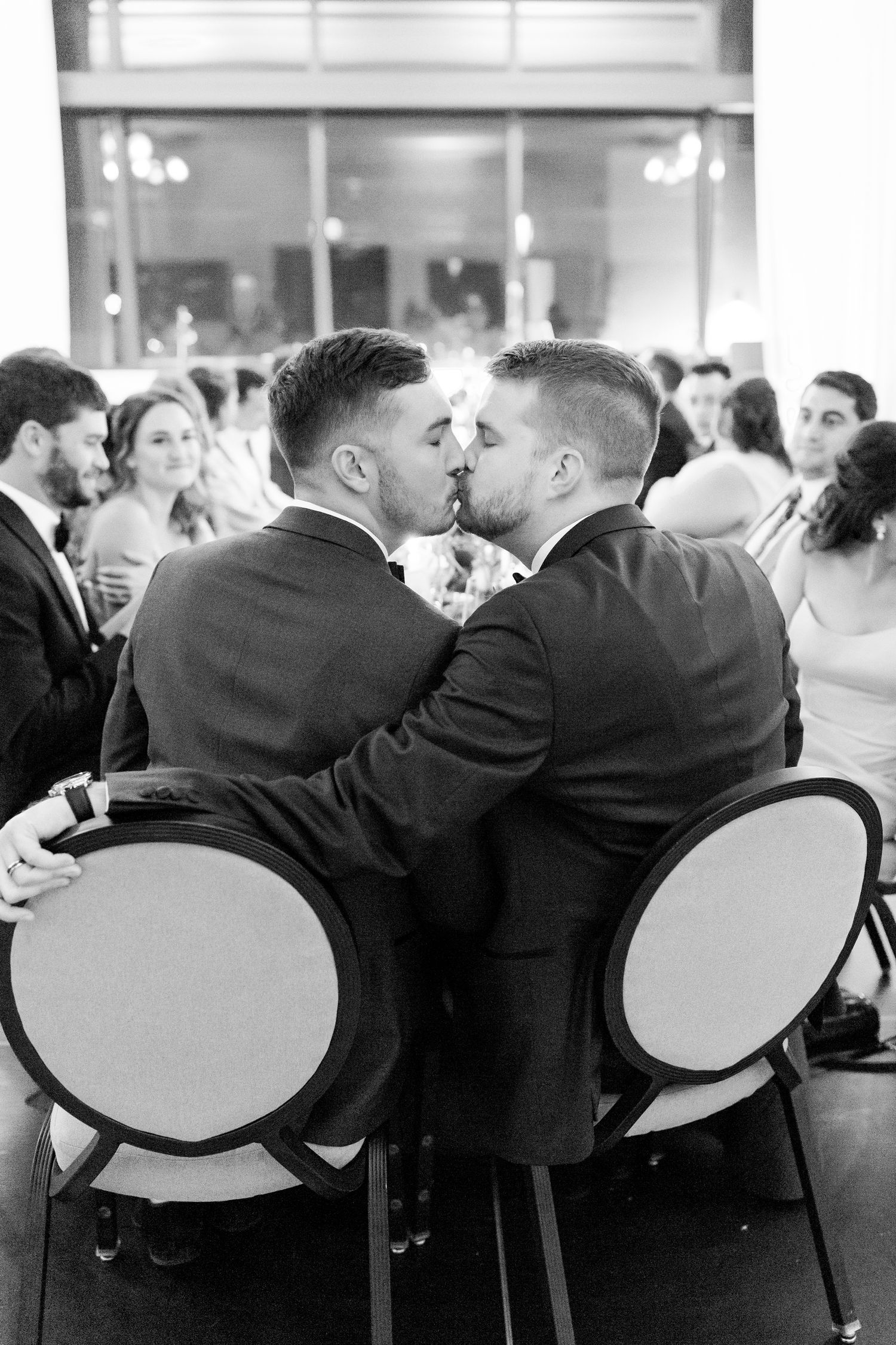 Two grooms in black suits share an intimate moment at their wedding reception while seated in elegant white-backed chairs.