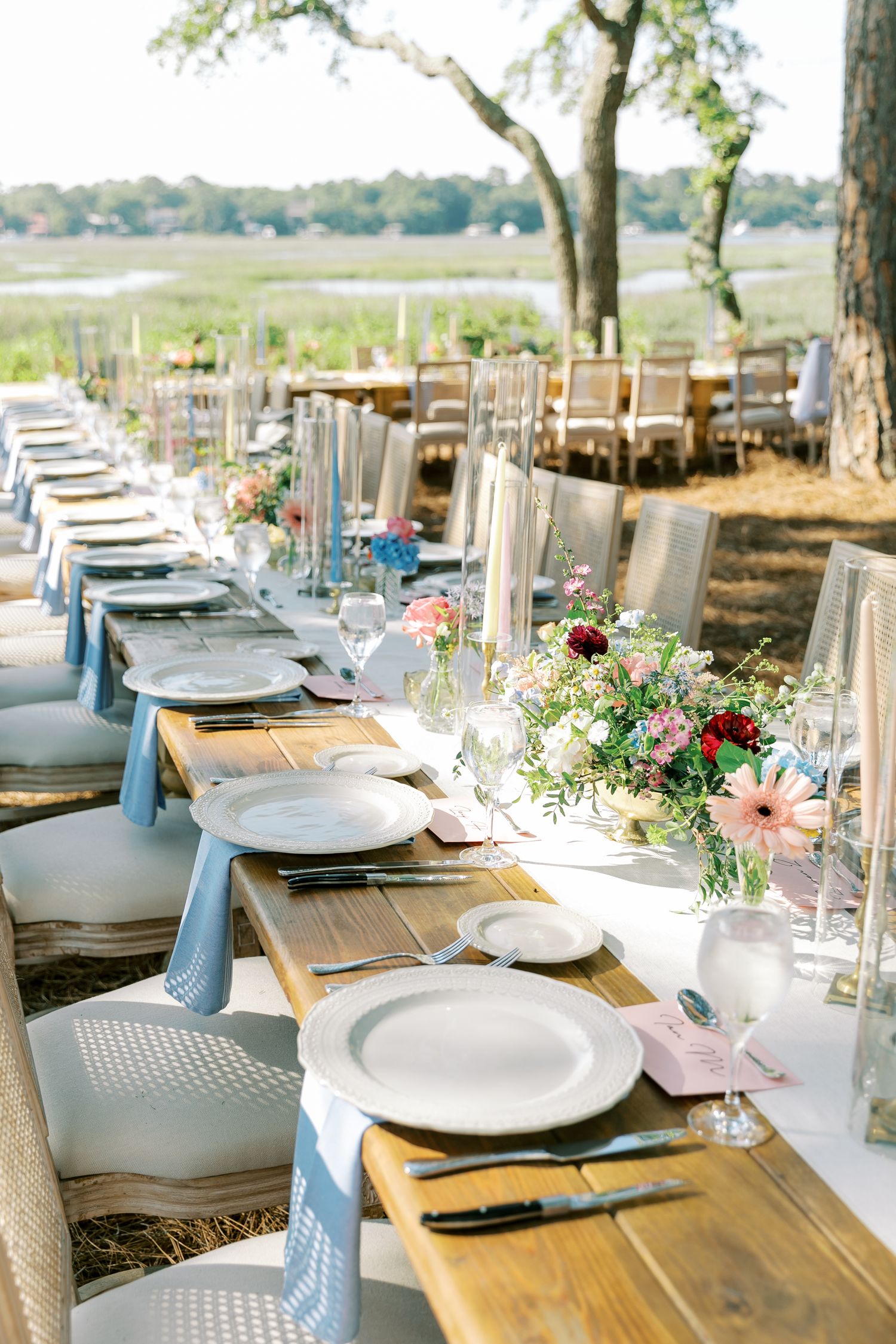 An elegant outdoor wedding table setting with rustic wooden tables and floral centerpieces overlooking a marsh.
