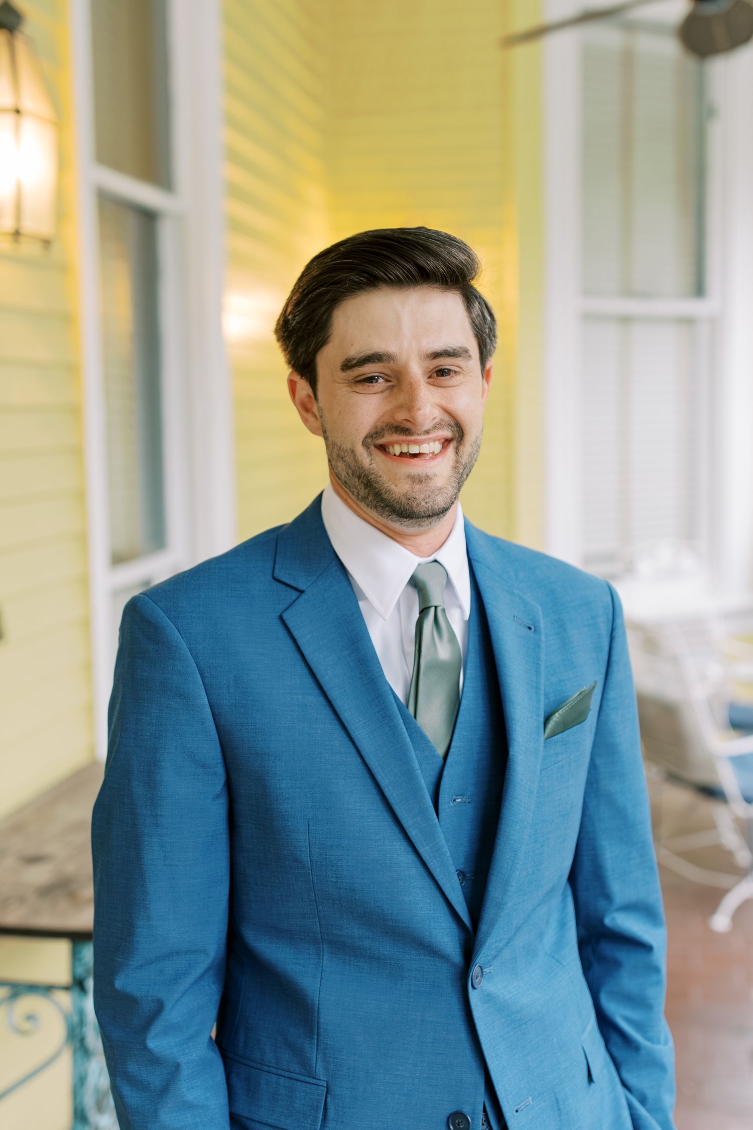 Groom wearing a stylish bright blue suit with gray tie poses on a yellow house porch.