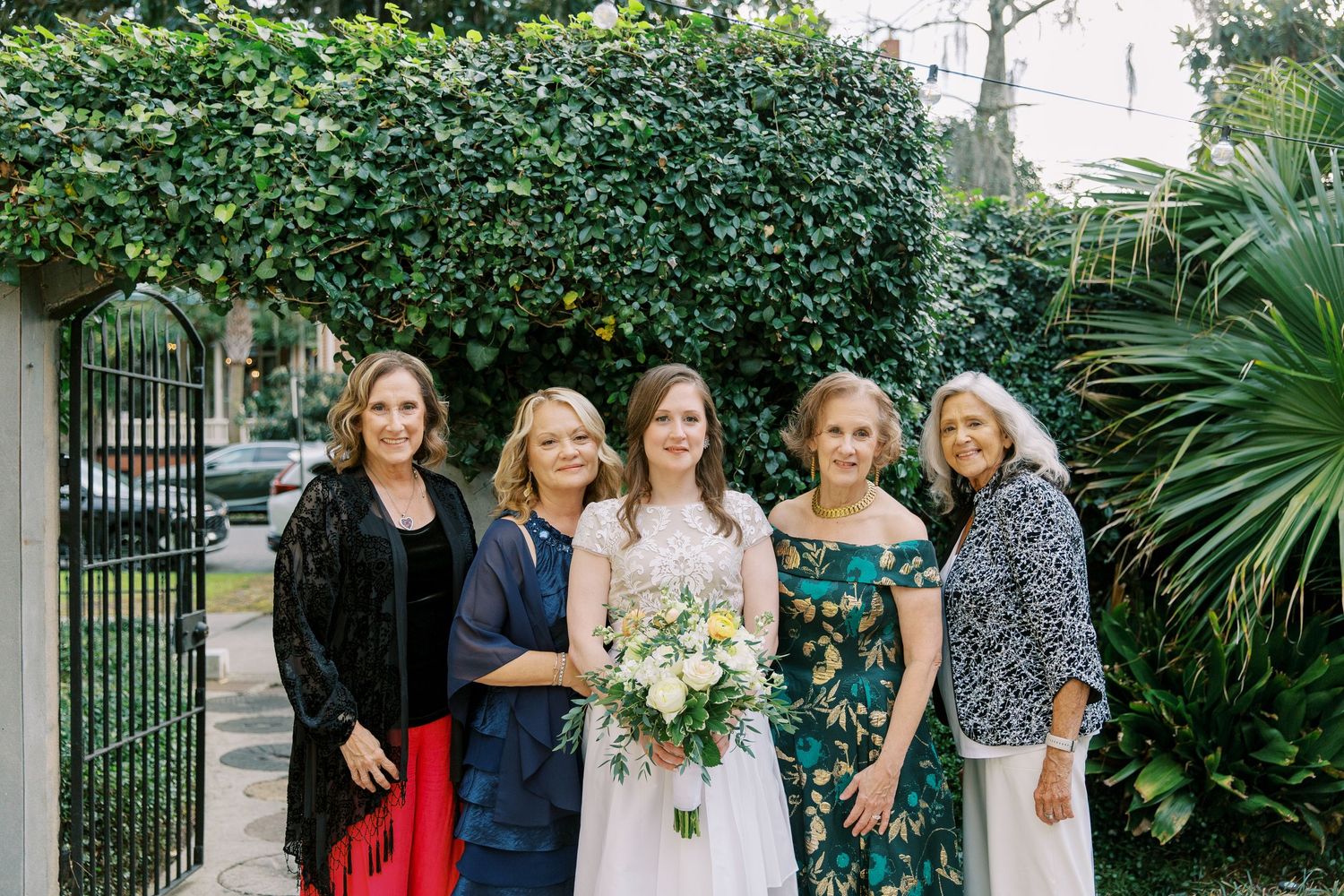 Wedding party poses together in a lush garden setting with greenery and palm trees in the background.