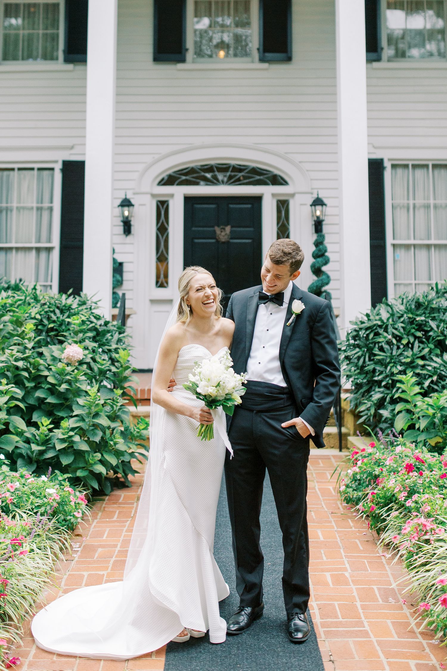 Classic wedding portrait in front of white colonial house with manicured garden path.