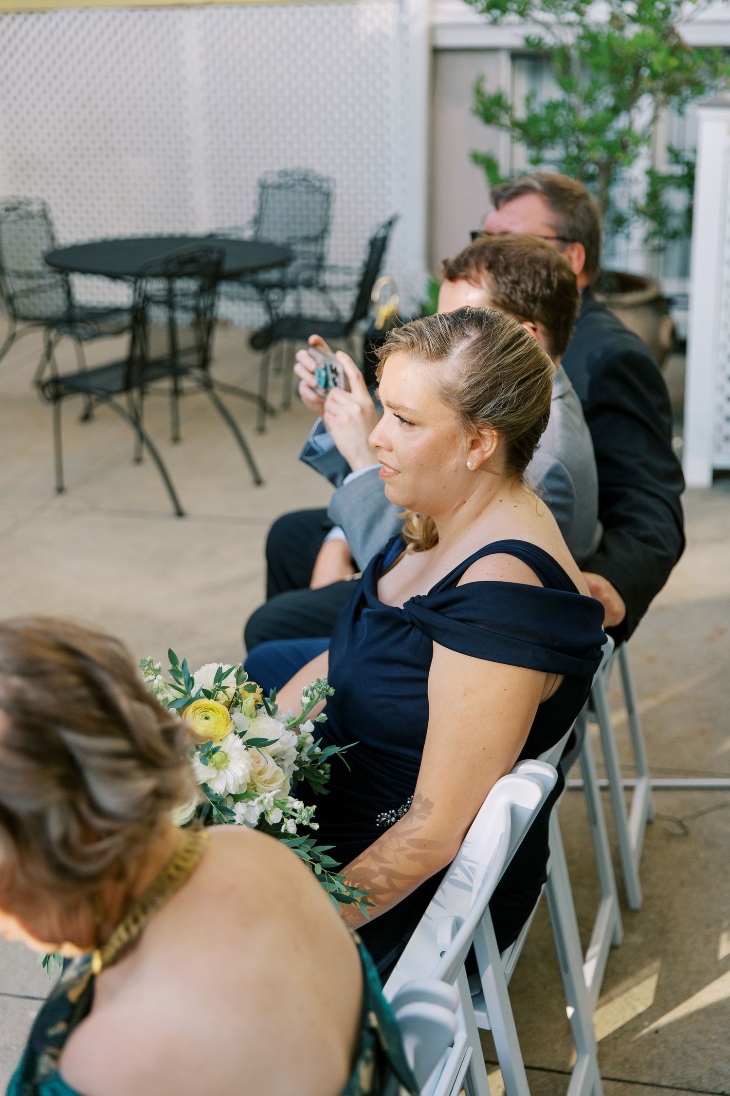 Wedding guests in dark attire sit in outdoor chairs at a ceremony.