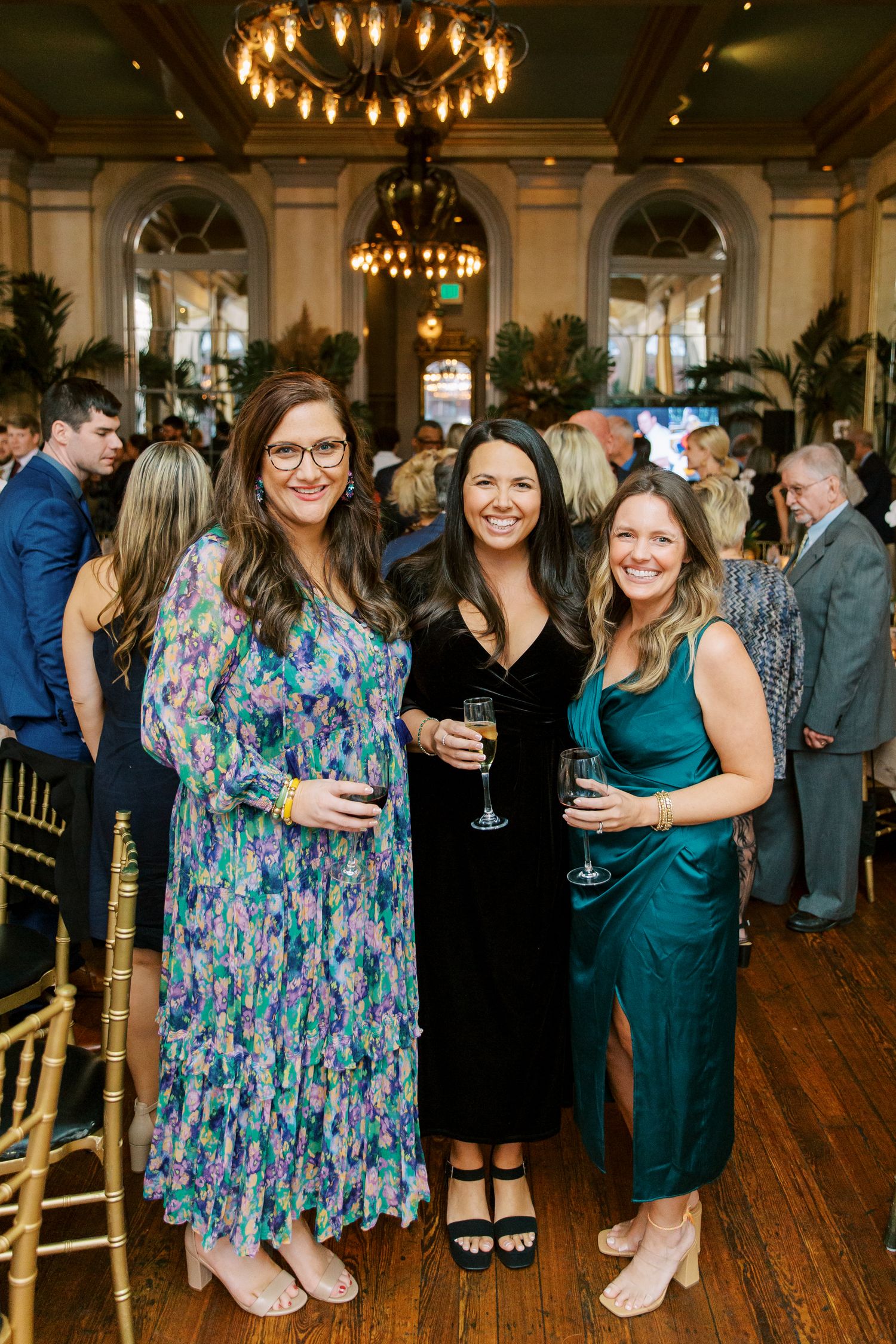 Three friends in formal attire gather at an elegant venue with chandeliers and hardwood floors during a social event.
