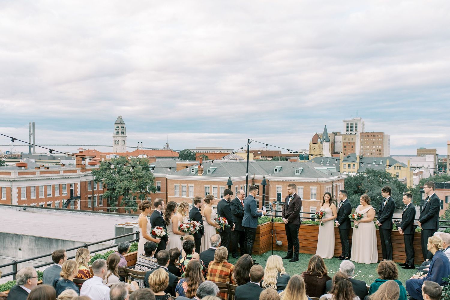 An outdoor rooftop wedding ceremony takes place against a cityscape backdrop with historic buildings and cloudy skies at Perry Lane Hotel