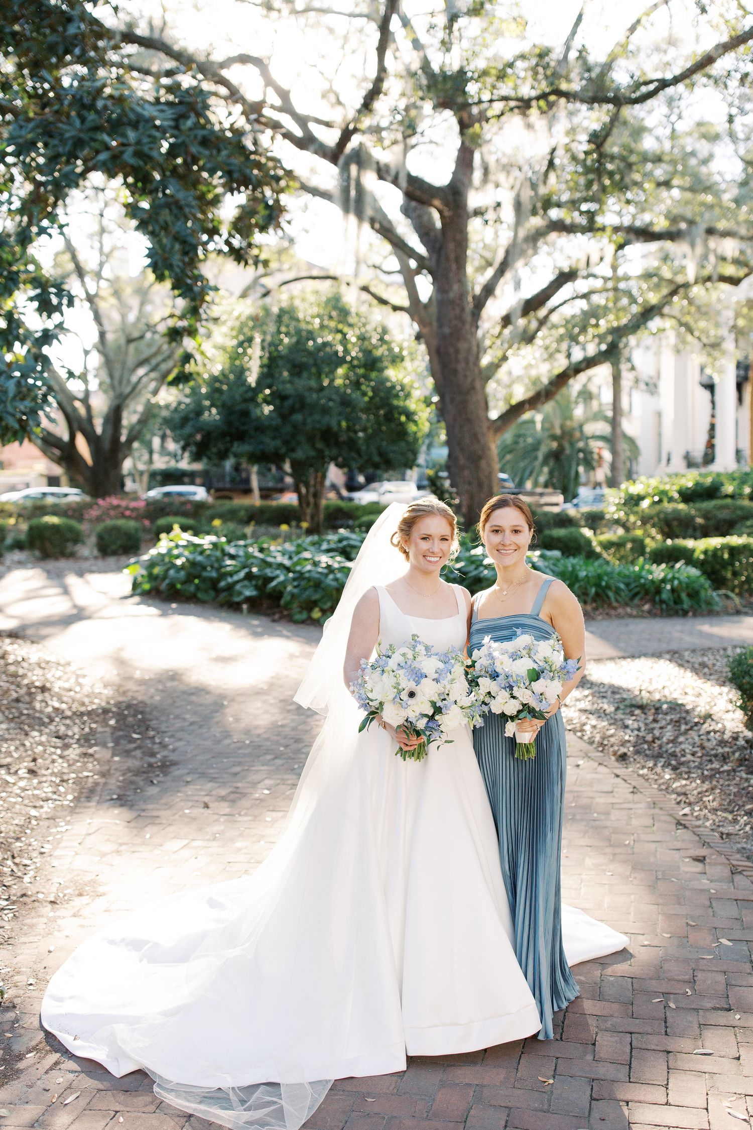 Wedding party with blue bridesmaid dress poses on brick pathway under oak trees.