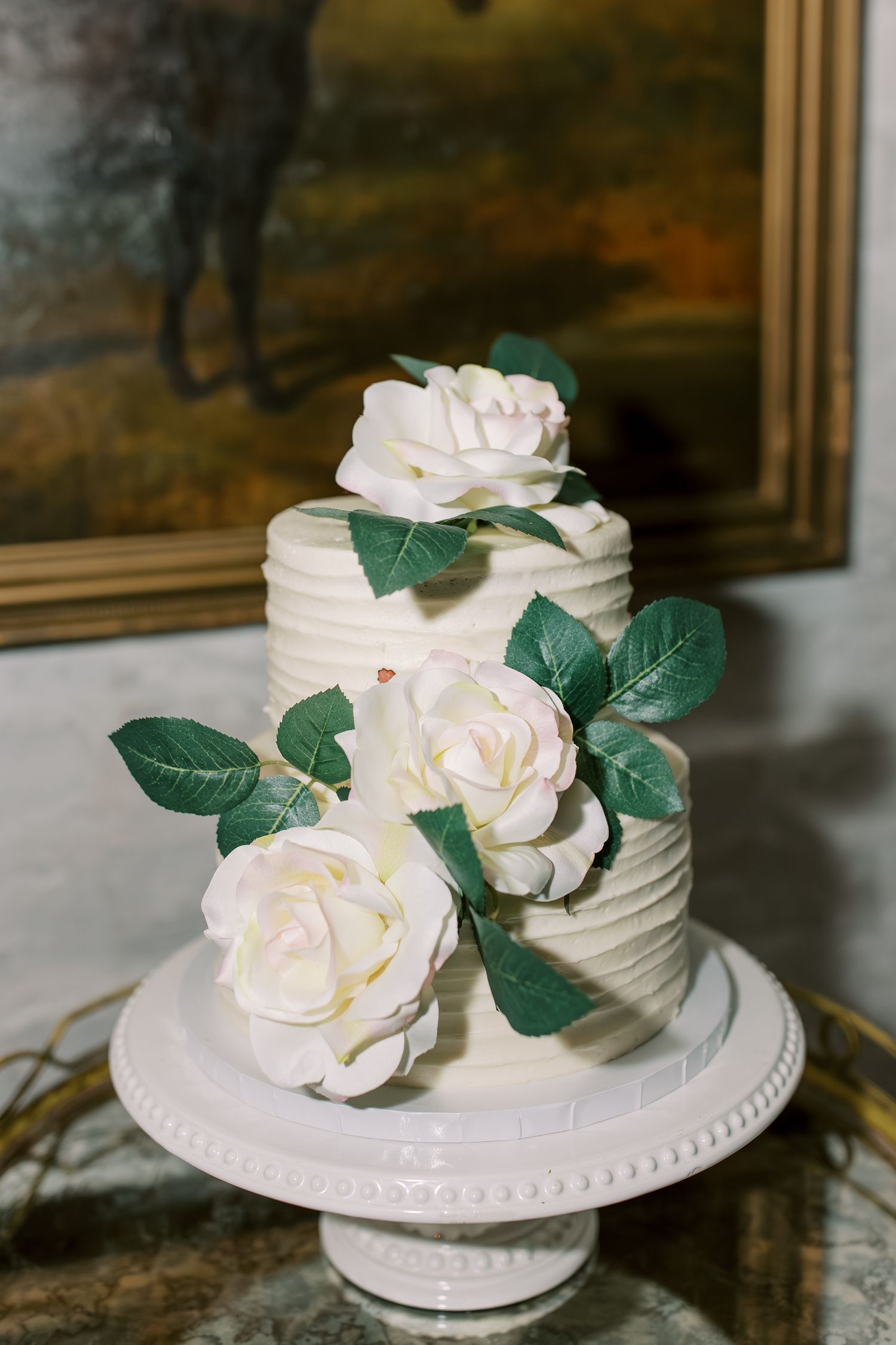 An elegant white wedding cake decorated with realistic sugar roses and green leaves on a vintage cake stand.
