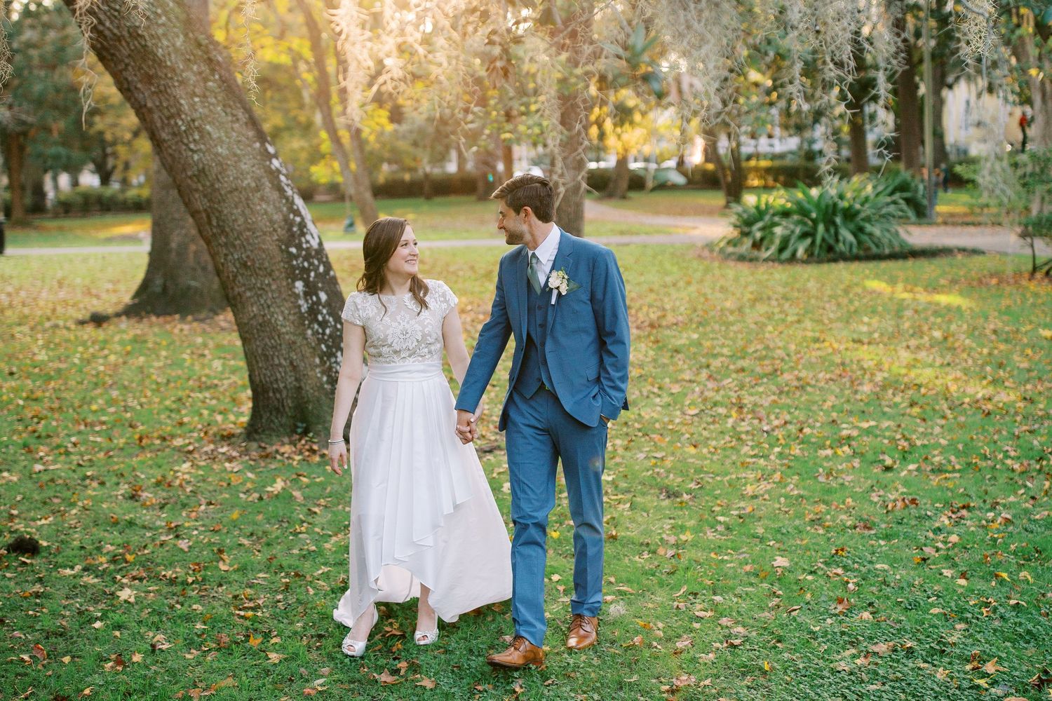 Two people in wedding attire walk together through a park filled with autumn leaves on a sunny afternoon.