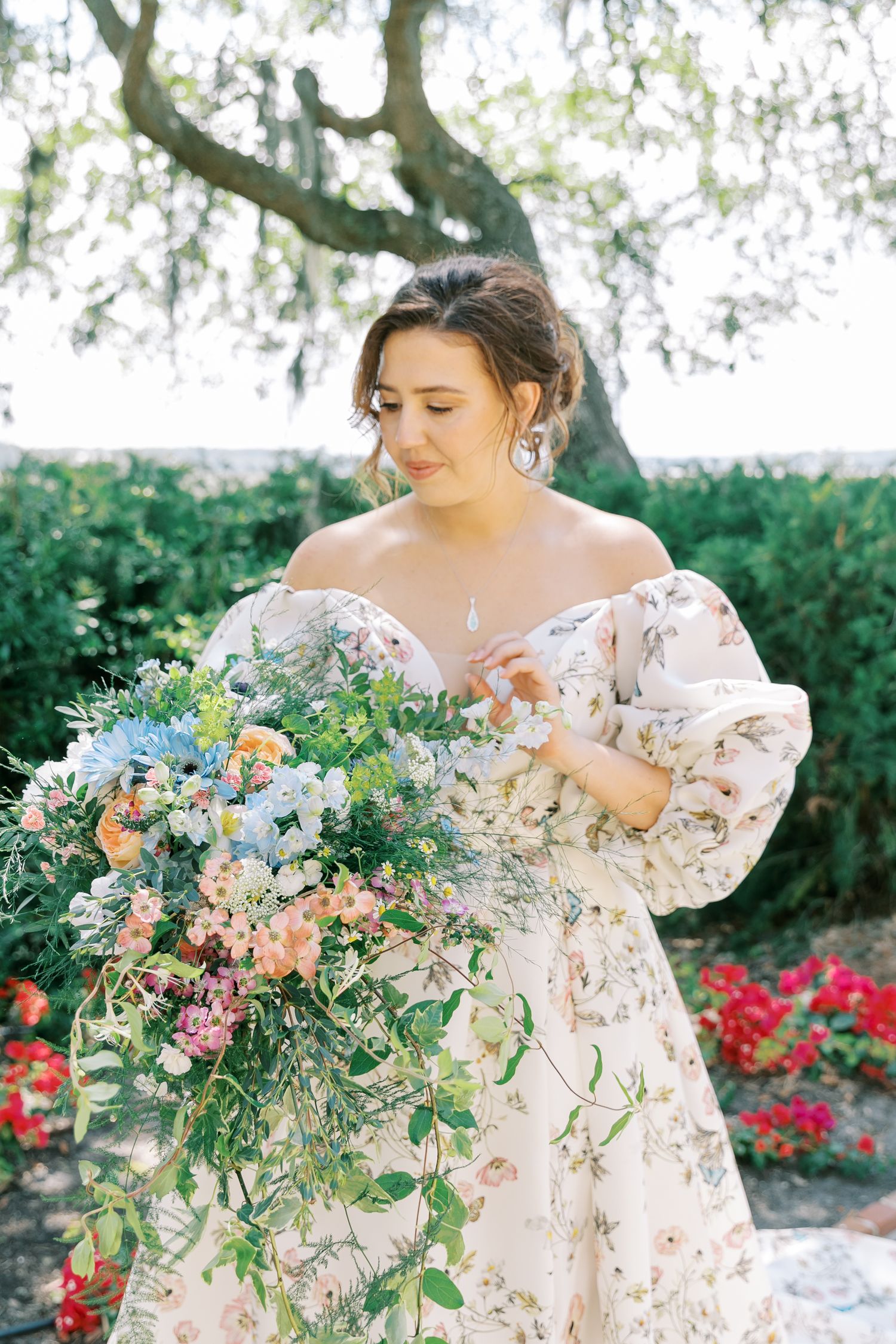 Bride in a floral off-shoulder gown holding a lush bouquet of pastel flowers in a garden setting.