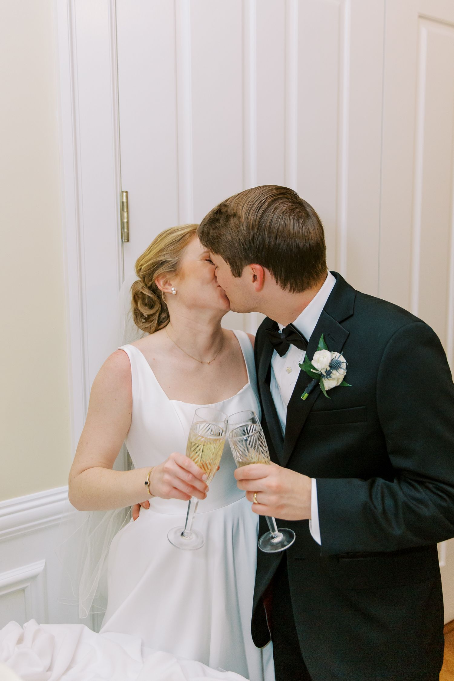 Romantic wedding couple sharing champagne toast in elegant white interior setting.